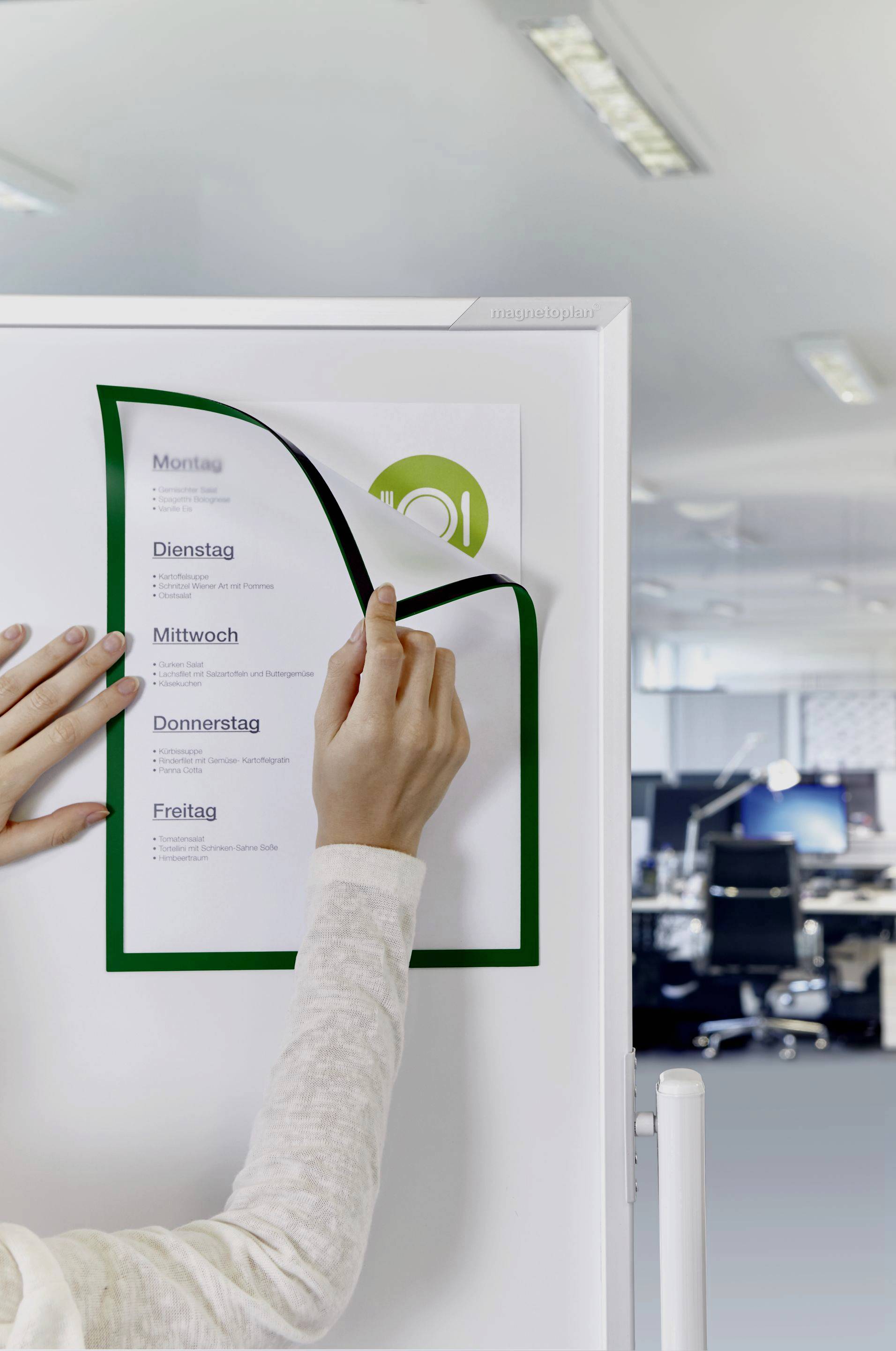 Une personne accroche une feuille avec des jours de la semaine sur un tableau de bureau. Des meubles de bureau sont flous en arrière-plan.