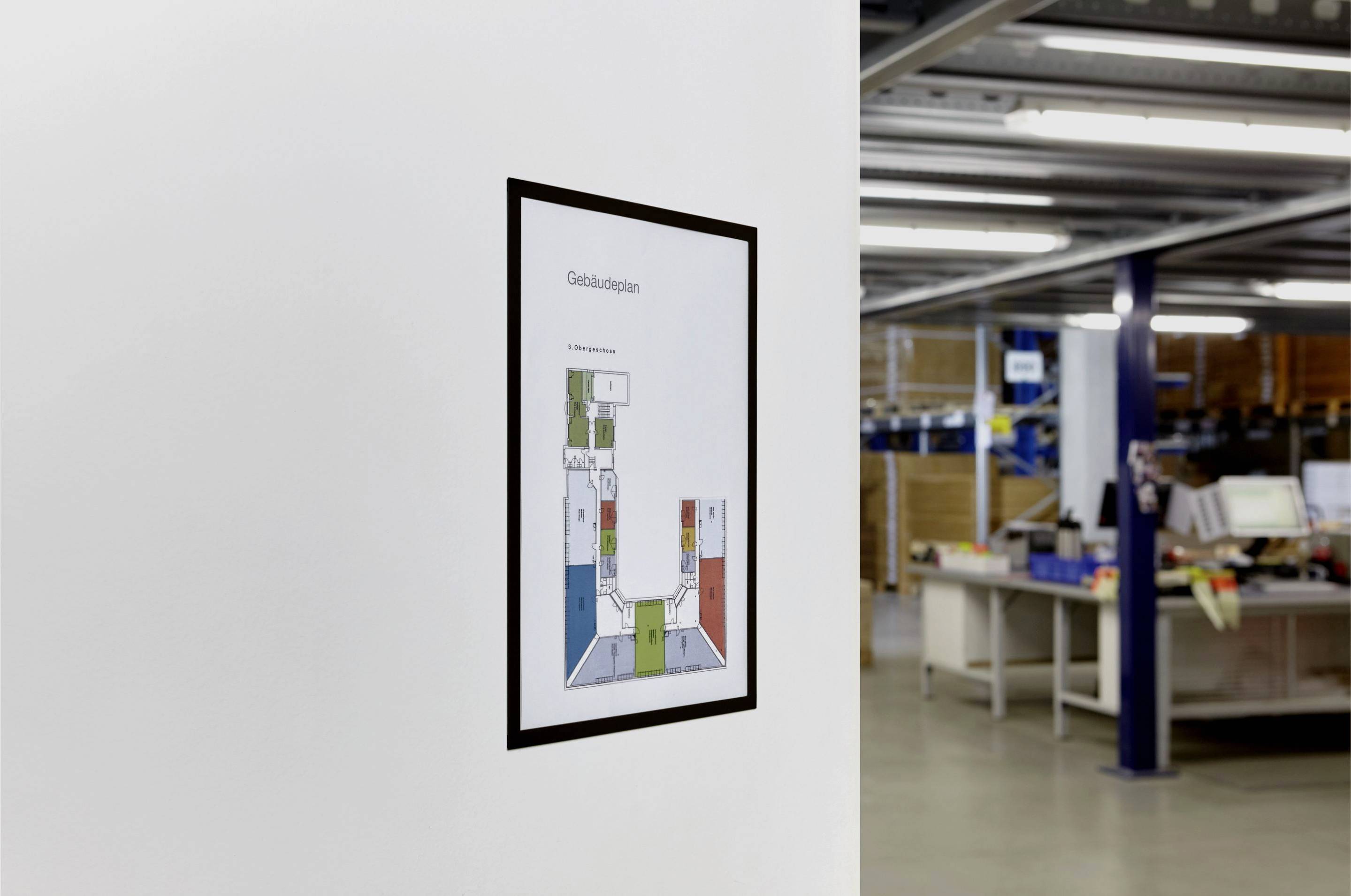 Un panneau indicateur est accroché à un mur blanc dans un bureau ou un entrepôt. Des étagères et des bureaux sont visibles en arrière-plan.