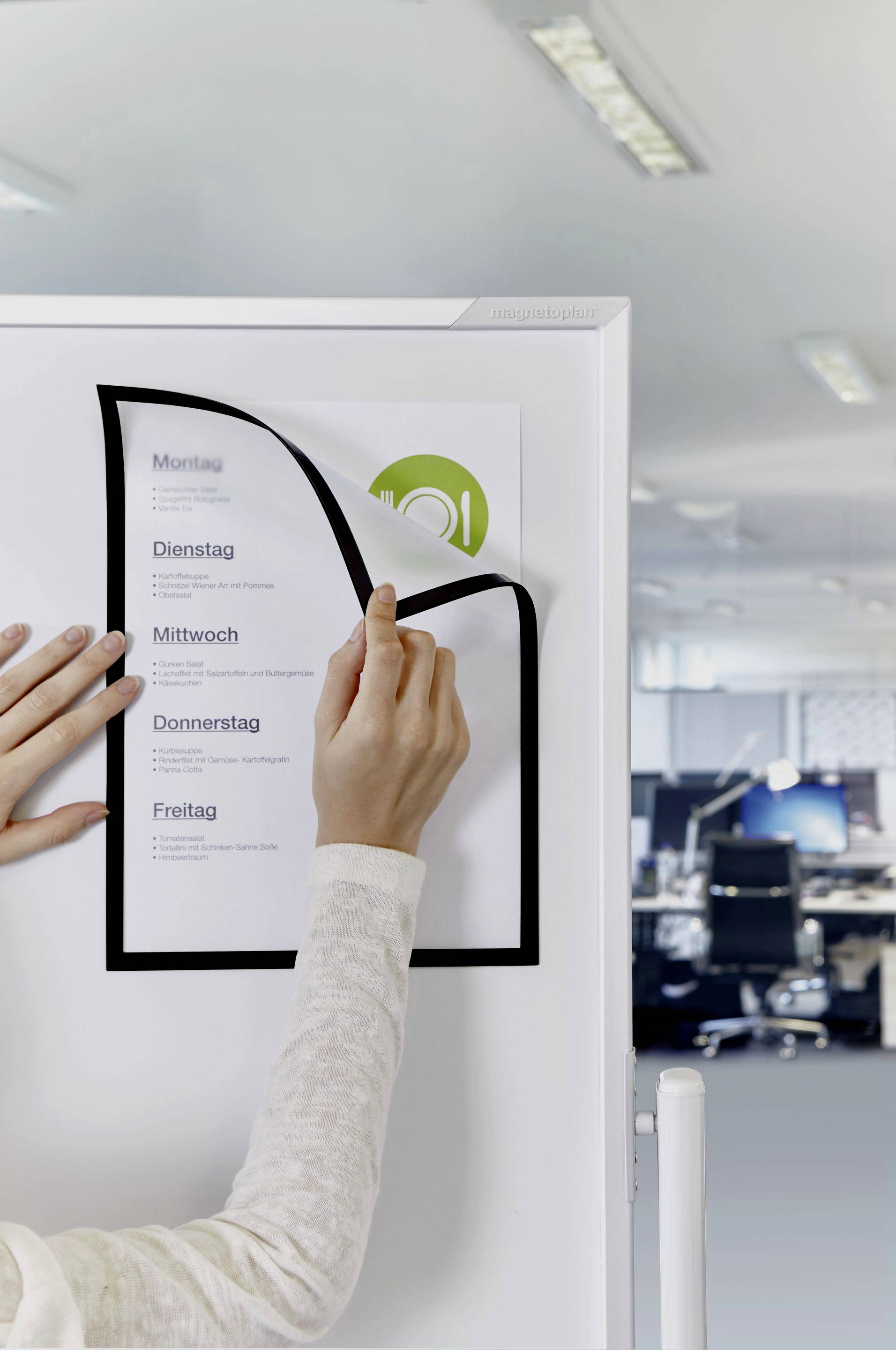 Une personne colle une feuille avec une liste des jours de la semaine (de lundi à vendredi) sur un tableau dans un bureau moderne.