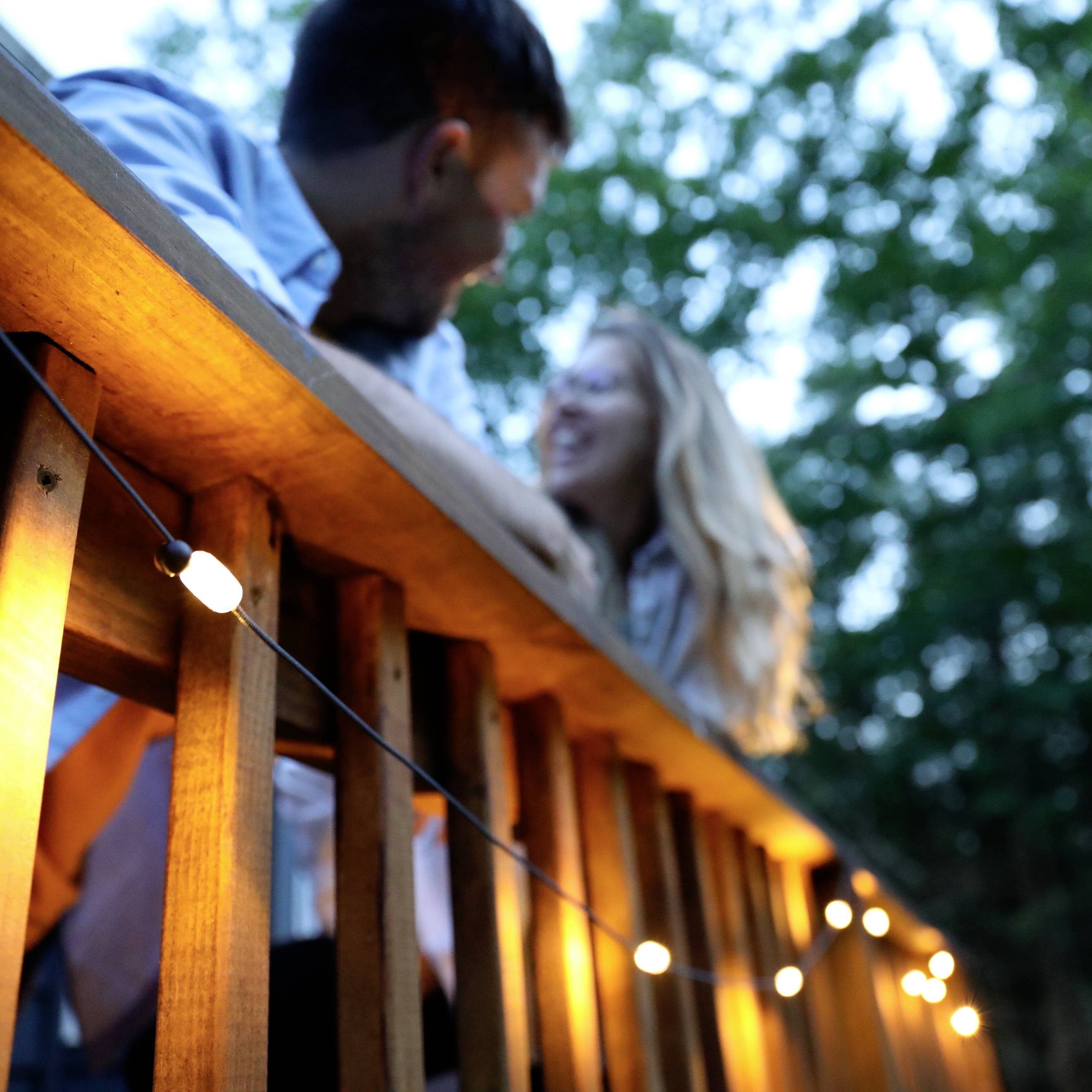 Un couple se regarde tendrement dans les yeux sur une terrasse décorée de guirlandes lumineuses, entouré d'arbres verdoyants.