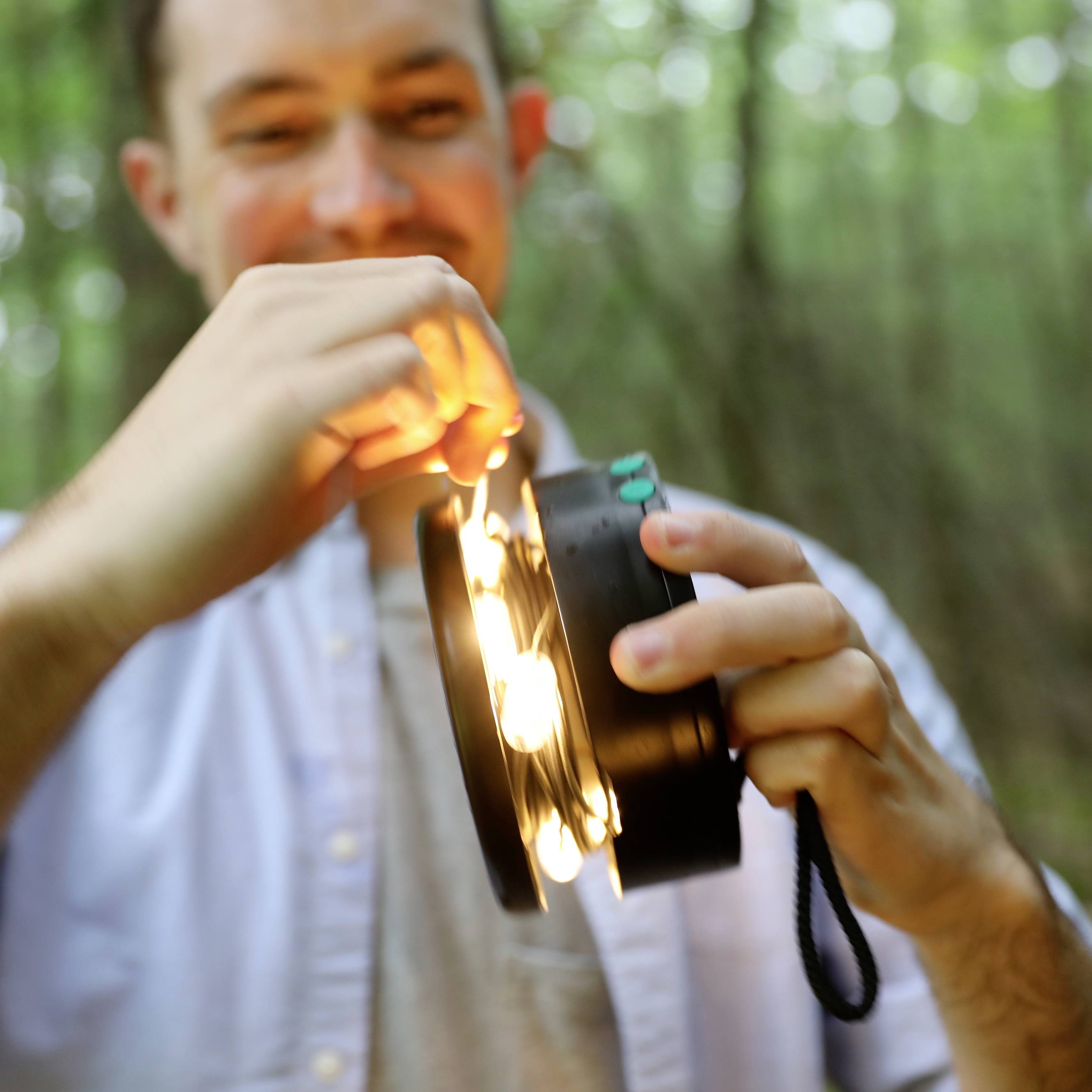 Un homme à l'extérieur tient une lampe ronde avec des ampoules lumineuses à la main. En arrière-plan, on aperçoit une forêt.
