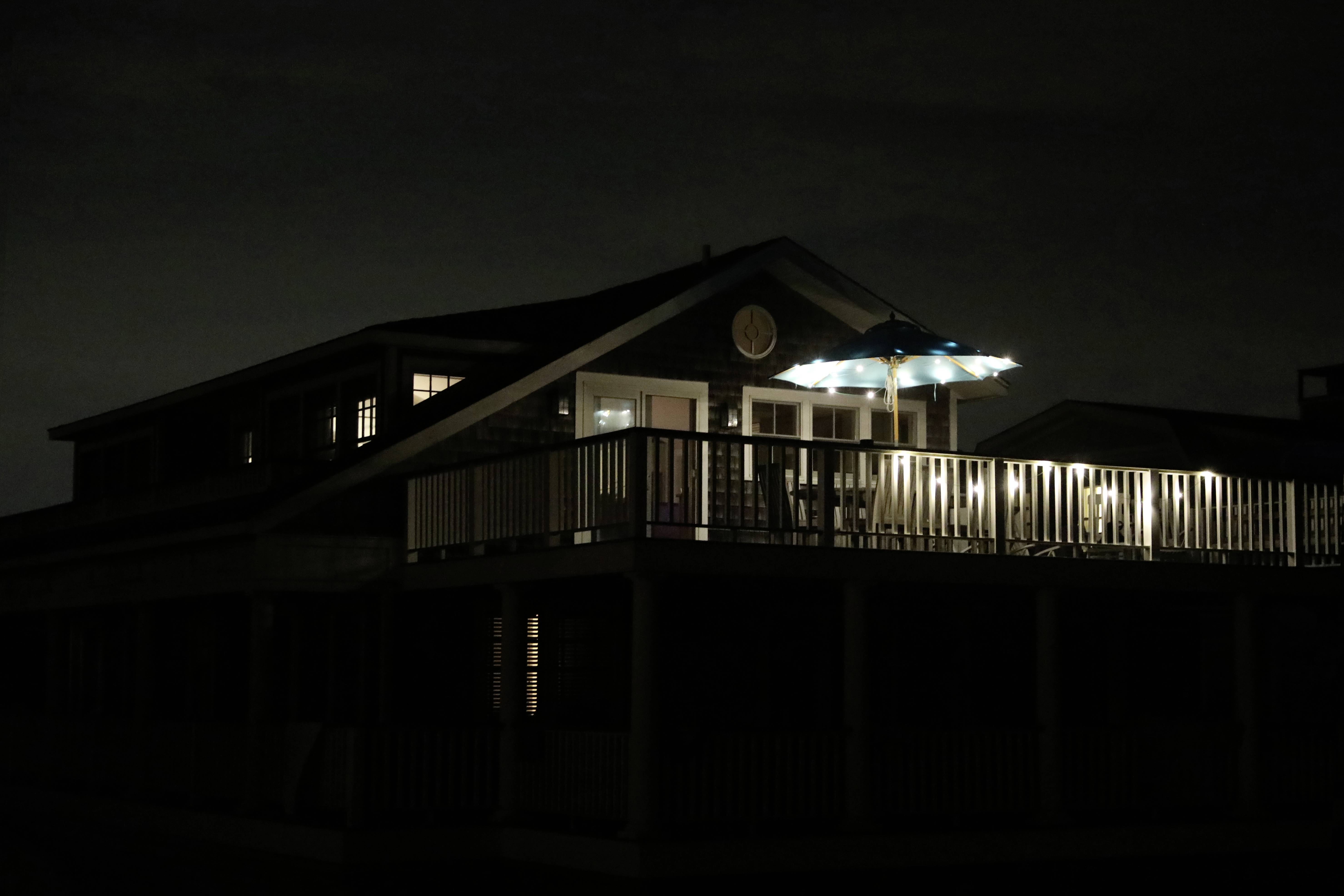 Une maison la nuit, faiblement éclairée par des fenêtres et un parasol lumineux sur le balcon, transmet une atmosphère paisible.