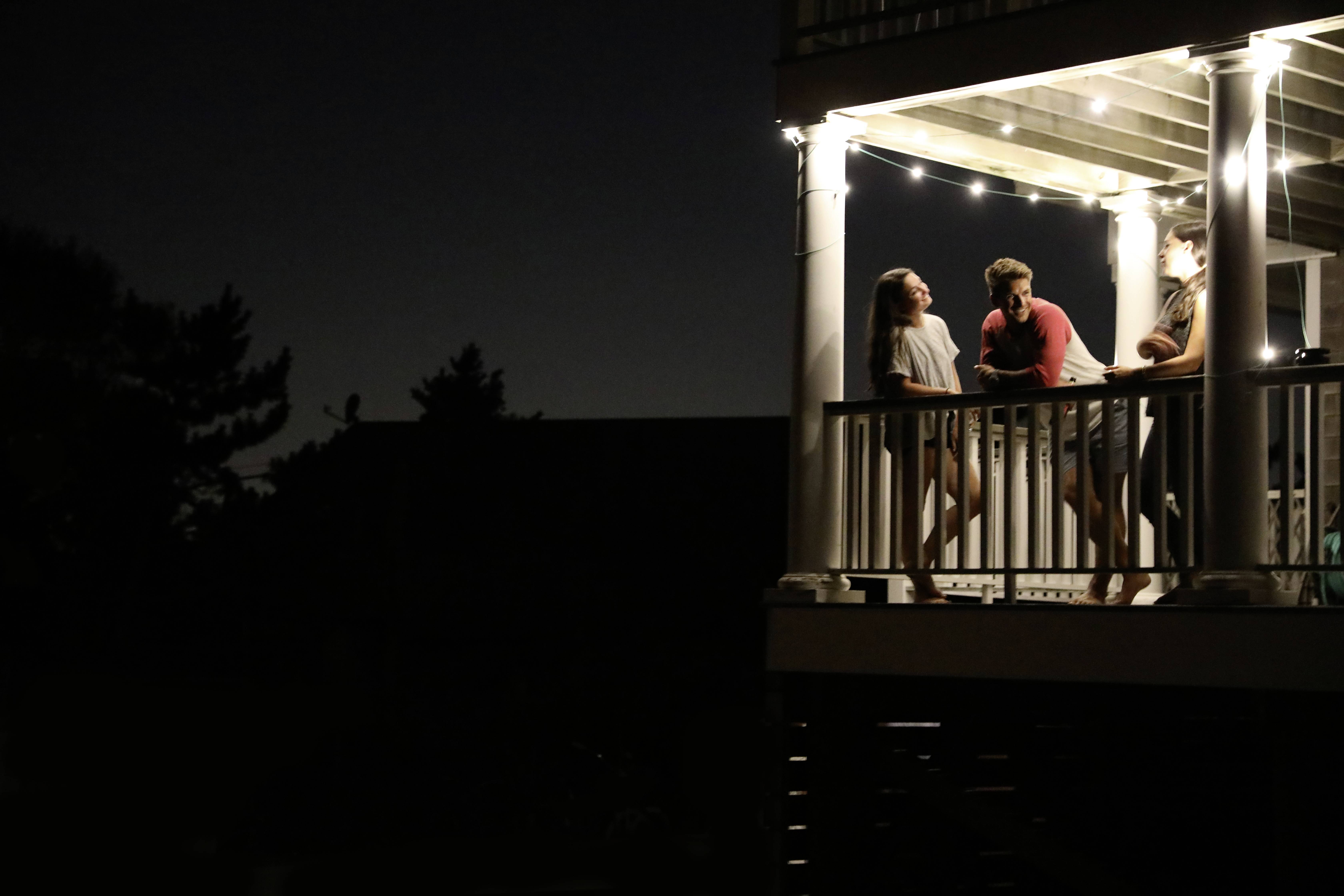 Quatre personnes discutent sur un balcon la nuit, des guirlandes lumineuses créant une ambiance chaleureuse et conviviale.