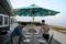 Deux personnes dressent une table à manger sous un parasol turquoise sur une terrasse donnant sur la plage.