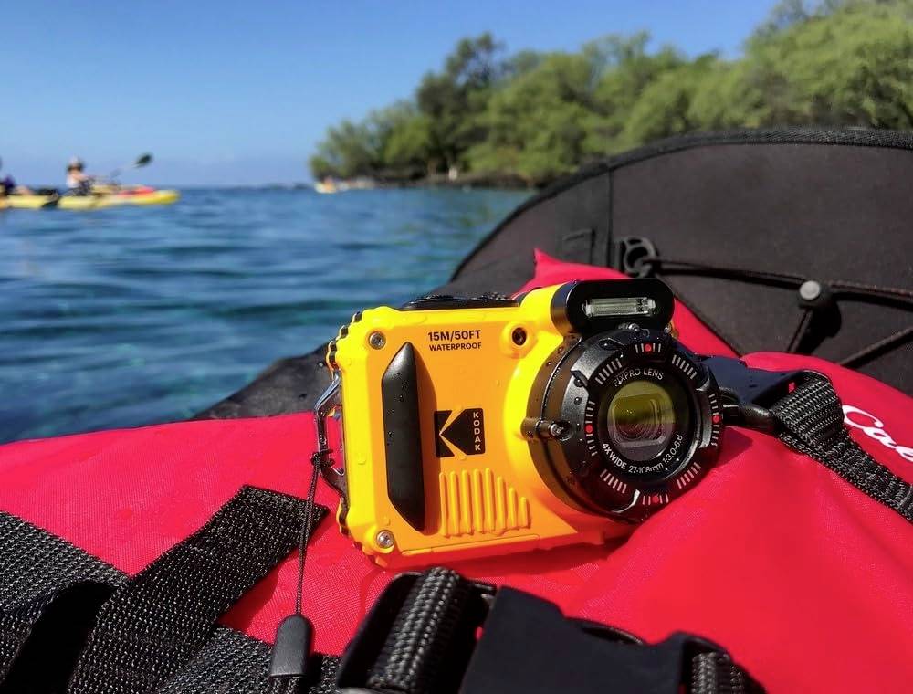 Un appareil photo jaune étanche est posé sur un gilet de sauvetage rouge dans un canoë, avec de l'eau bleue et des arbres verts en arrière-plan.
