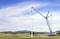 Une grande grue bleue construit une éolienne dans un vaste paysage. La grue soulève un composant, tandis que des montagnes sont visibles en arrière-plan.