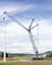 Un grand grue bleue soulève une partie d'une éolienne dans un champ. À l'arrière-plan, on aperçoit des collines et un ciel nuageux.