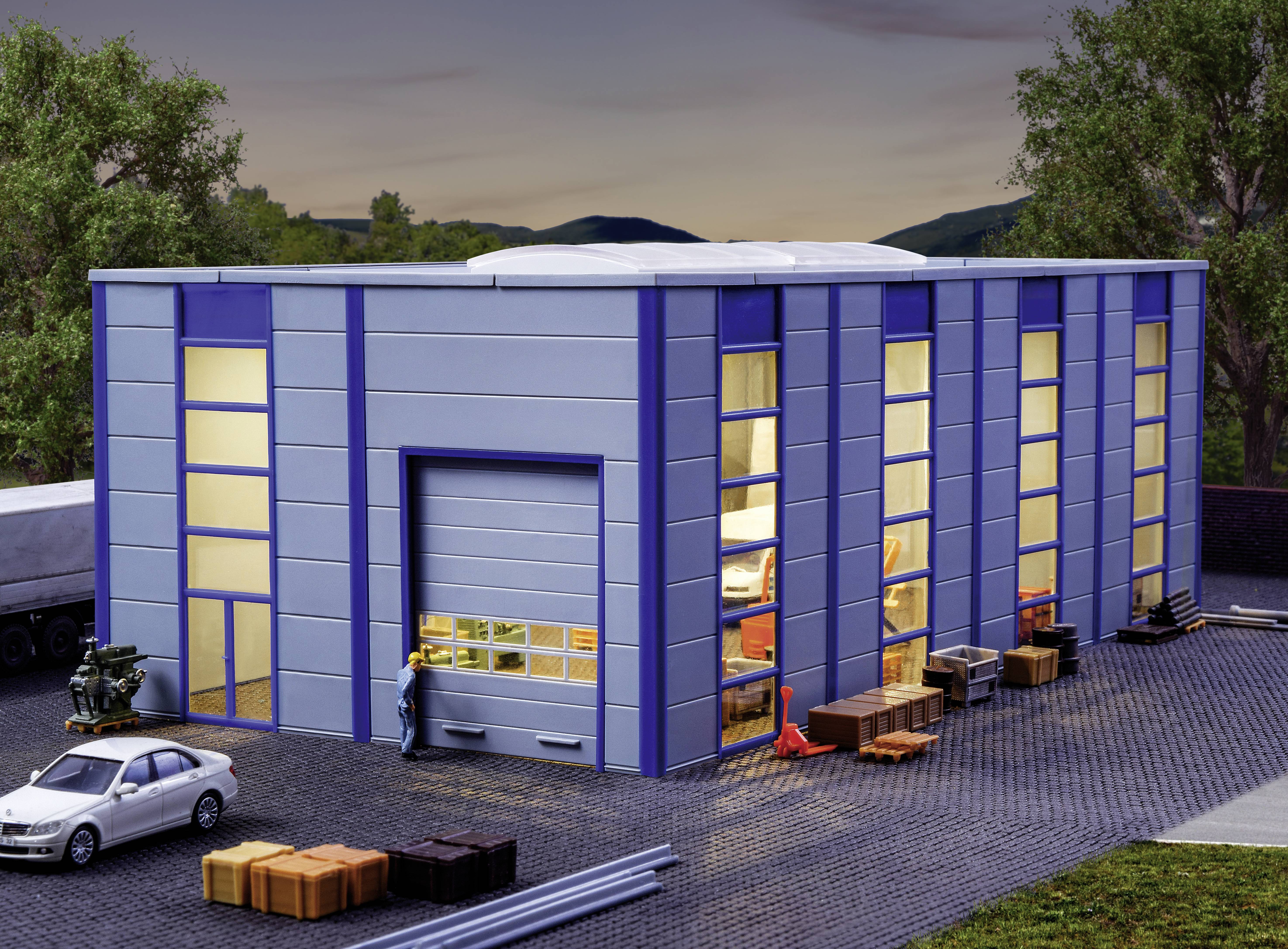 Un grand bâtiment industriel bleu avec plusieurs fenêtres, entouré d'arbres. Devant le bâtiment se trouvent une voiture et des objets empilés.