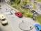 Rue modèle avec des miniatures de voitures, un passage piéton, une voiture rouge et un cycliste sur une piste cyclable le long d'un talus rocheux.