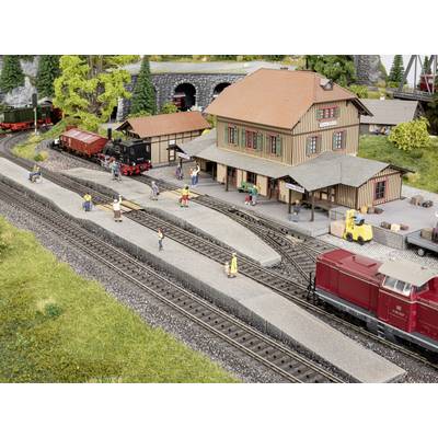 Scène de train miniature avec une petite gare. Des personnes attendent sur les quais. Trains sur les voies. Bâtiments à toits rouges en arrière-plan.