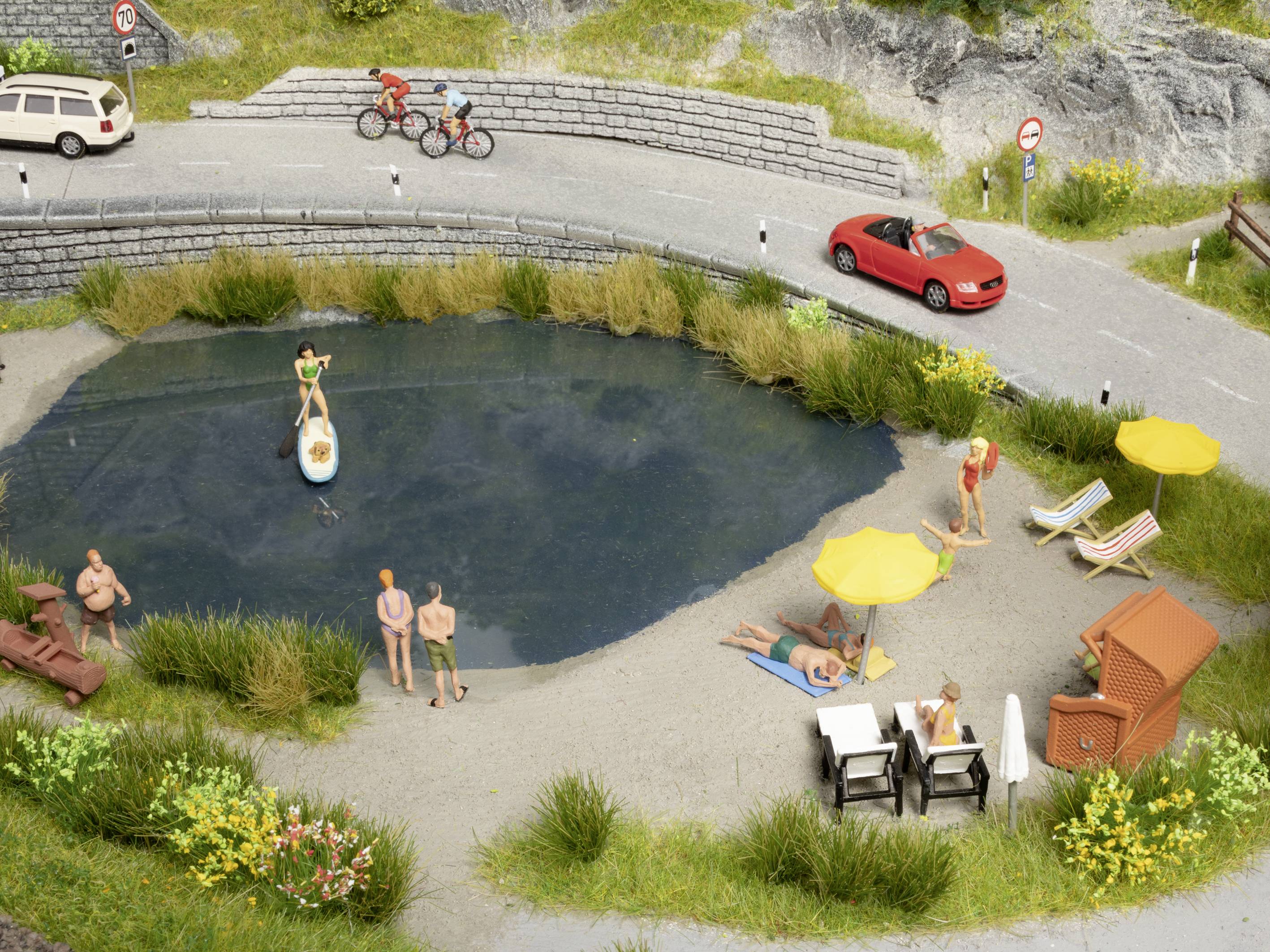 Un modèle miniature montre un petit lac avec une plage environnante. Des personnes se détendent au bord de l'eau, font du vélo et conduisent une voiture sur la route.