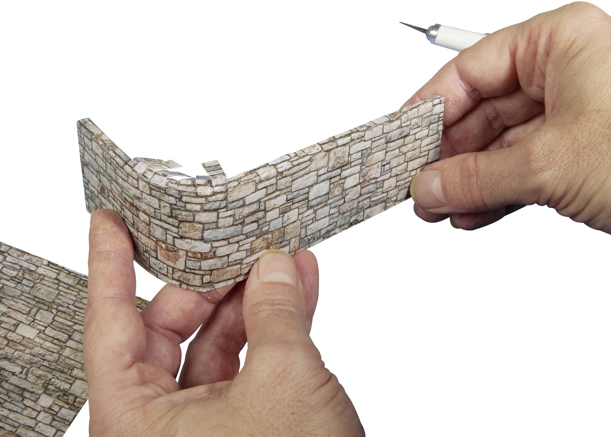 Deux mains forment une petite reproduction de mur en papier avec des pierres réalistes, probablement pour un projet de maquette.