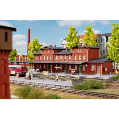 Une petite gare avec des bâtiments en briques rouges, entourée d'arbres verts et de petites figurines debout sur un quai.