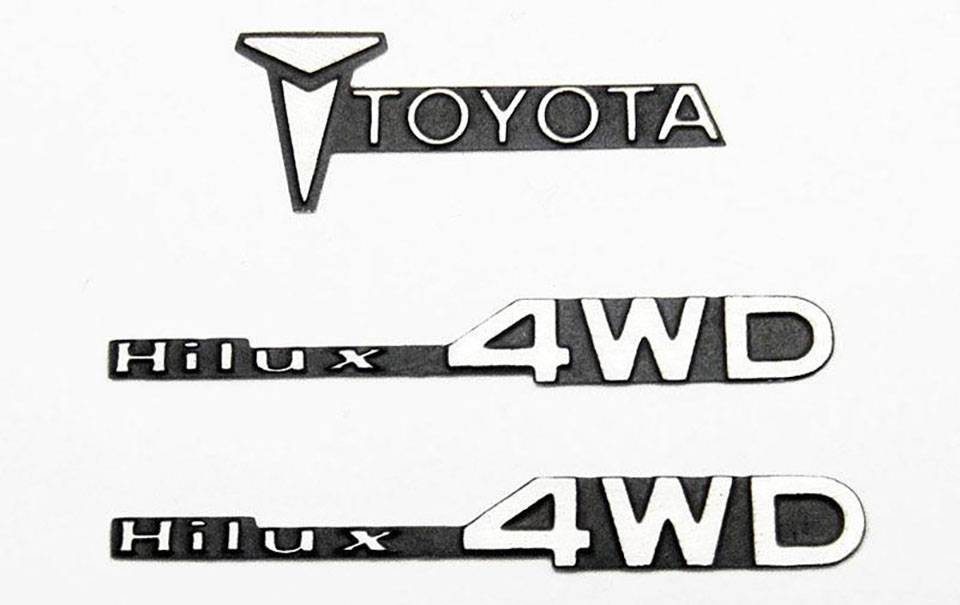 'Trois emblèmes : En haut le logo Toyota, en dessous deux fois le symbole Hilux 4WD. Tous en noir sur un fond blanc.'