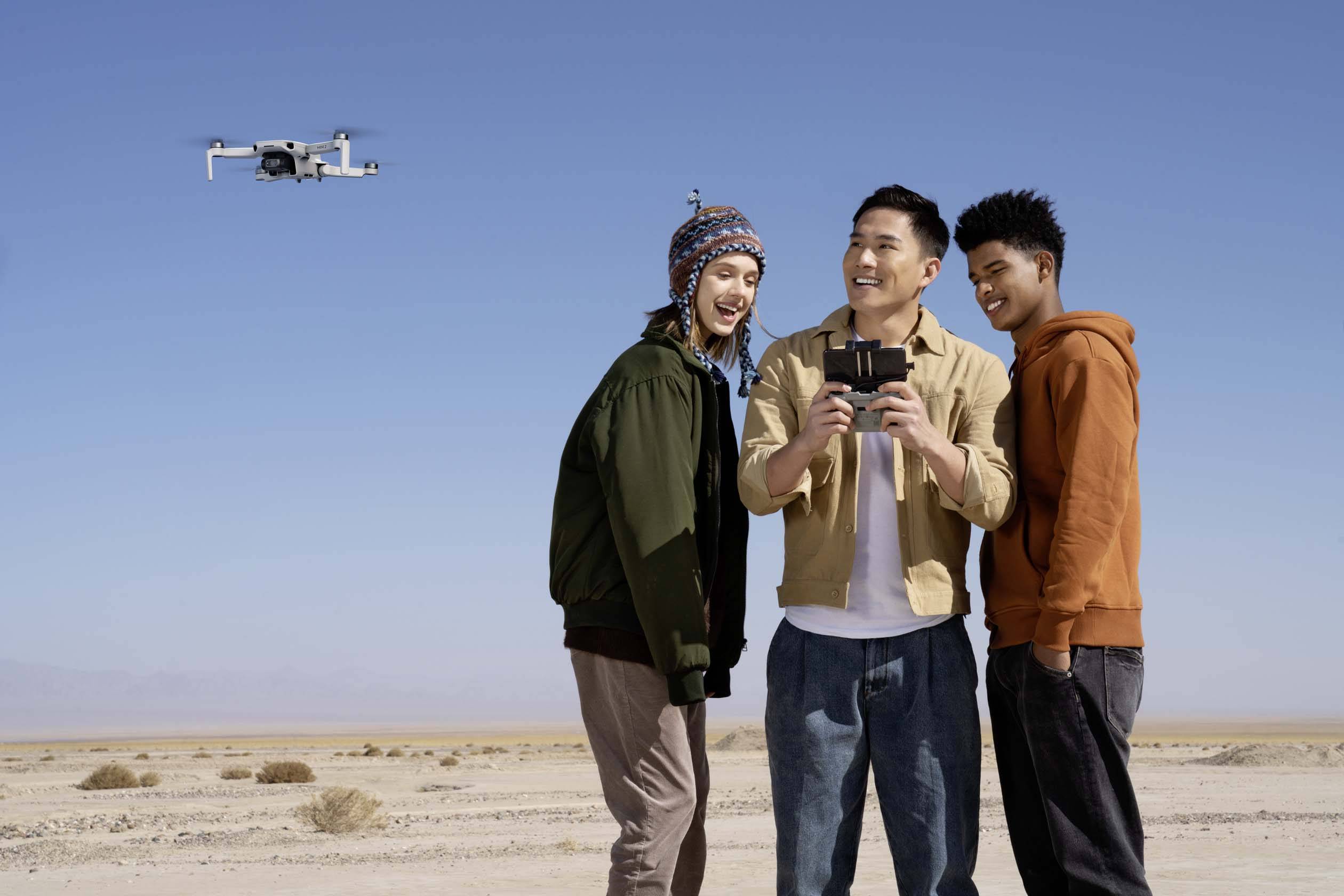 Trois personnes se tiennent souriantes dans le désert, regardant ensemble un smartphone. À l'arrière-plan, un drone survole le ciel bleu.