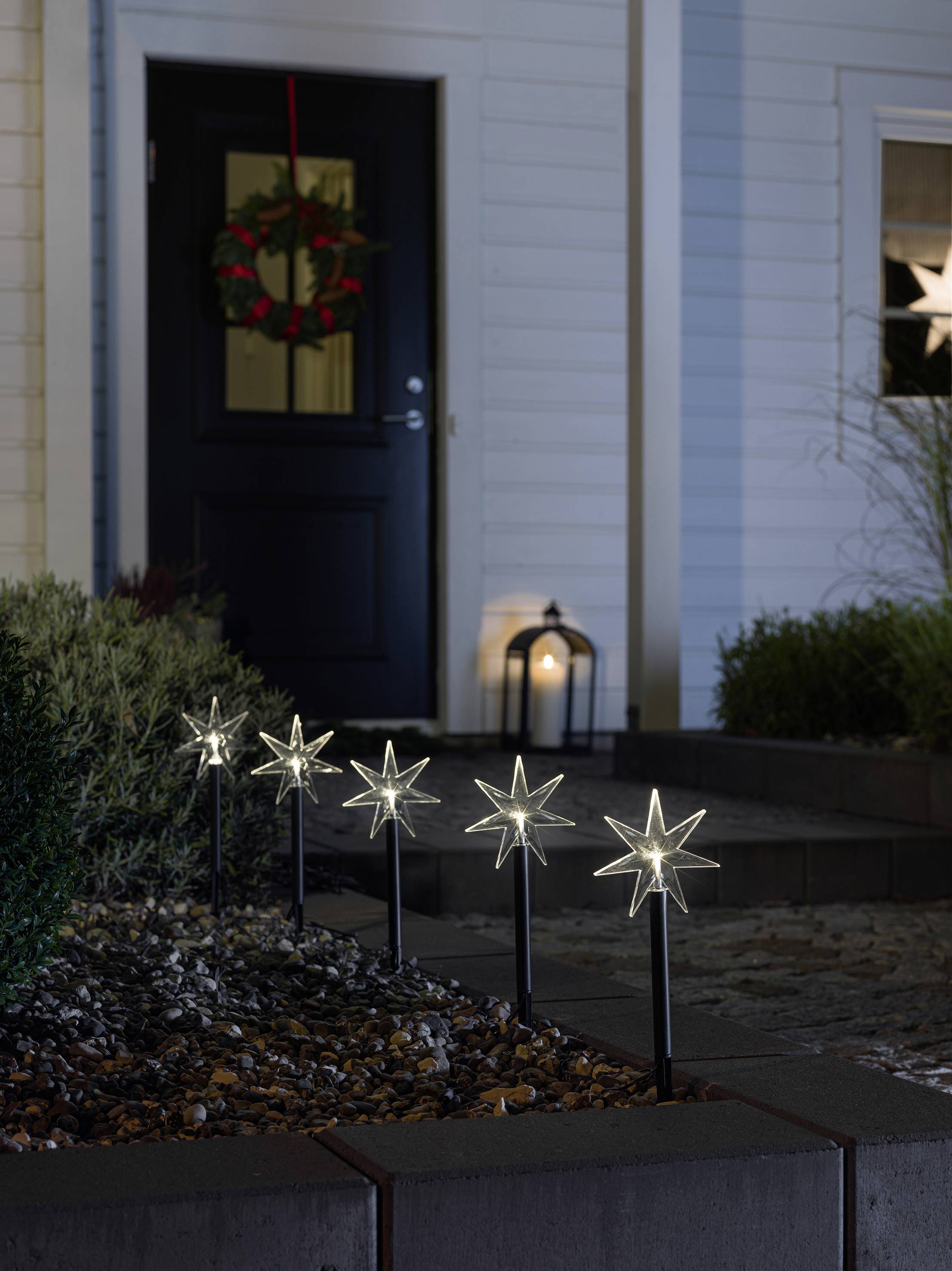 Un chemin de jardin devant une maison est bordé de lumières scintillantes en forme d'étoiles. Une lanterne et une couronne de Noël décorent la porte.