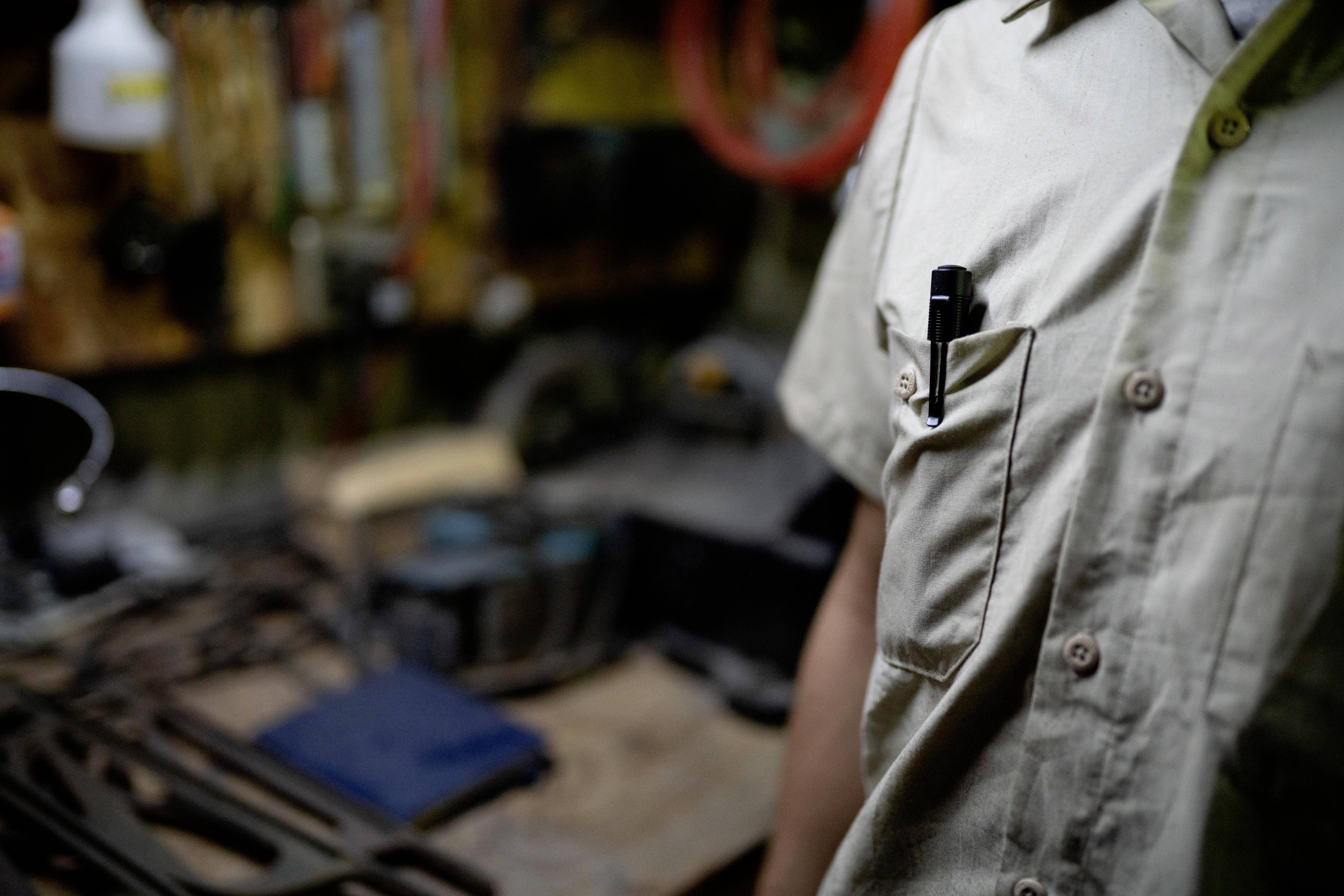 Un homme se tient dans un atelier rempli d'outils. Un stylo bille est glissé dans la poche de sa chemise. L'environnement est flou.