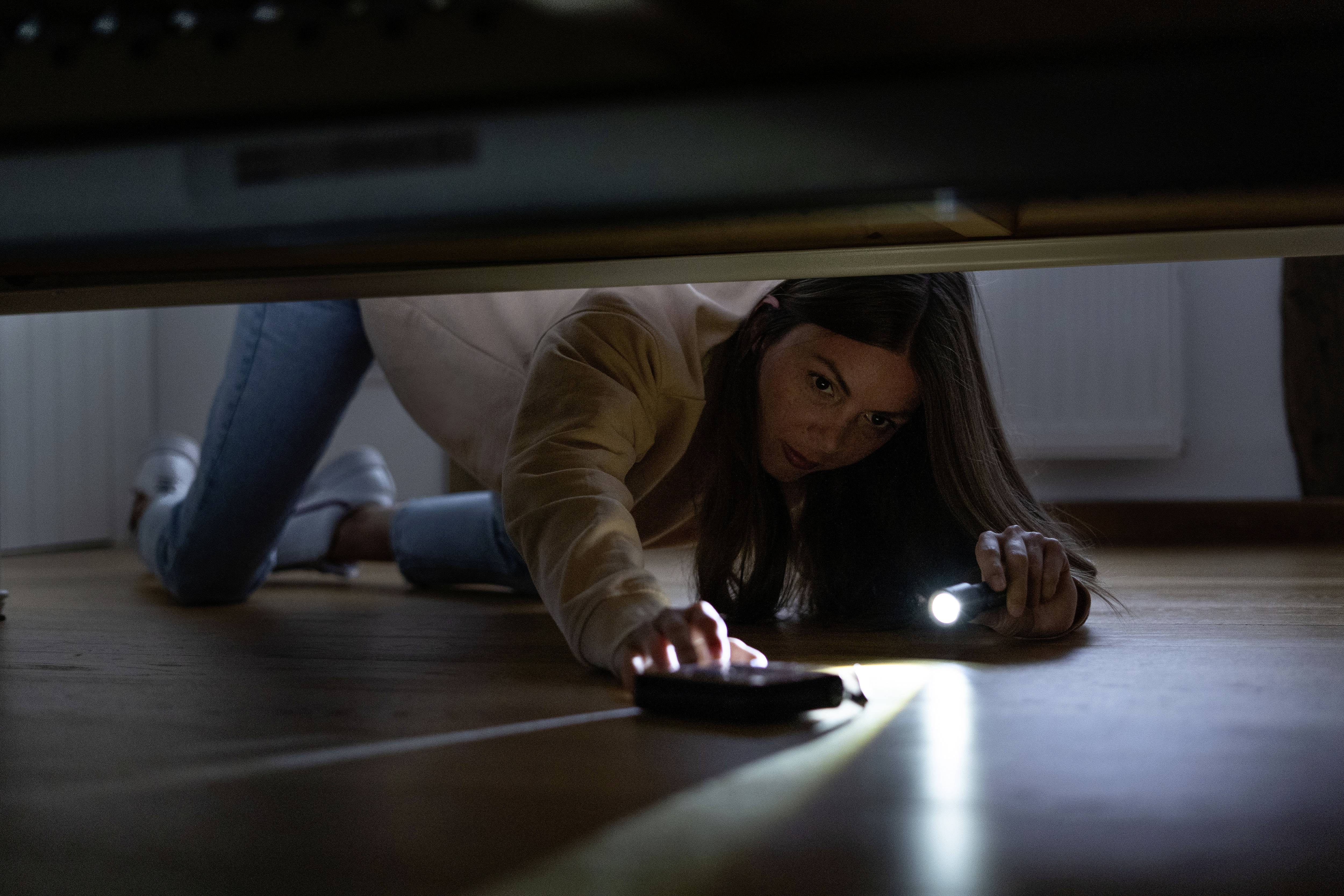 Une femme cherche son smartphone sous un canapé à l'aide d'une lampe de poche. Elle semble concentrée et déterminée dans sa recherche.