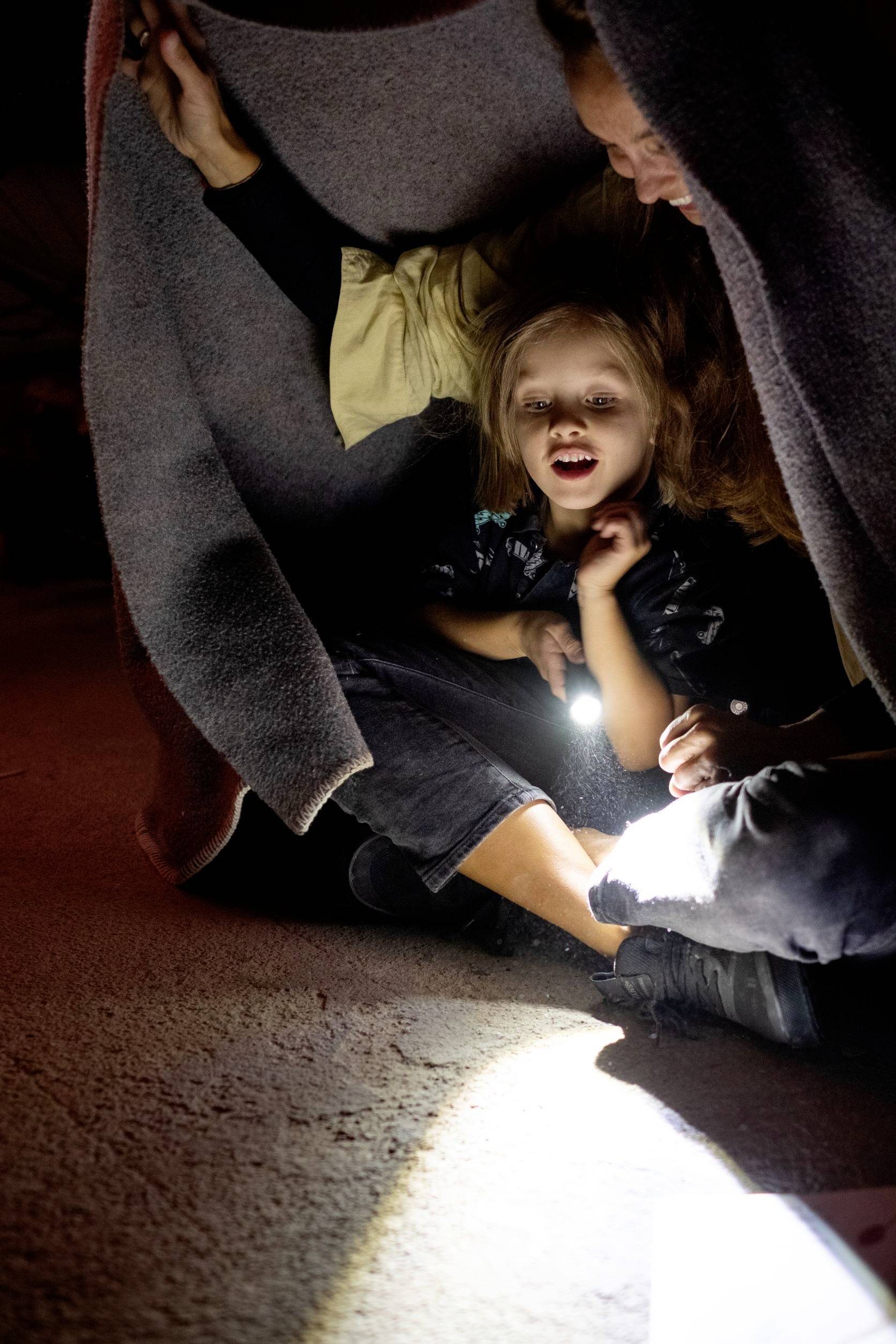 Un enfant et un adulte sont assis sous une couverture, l'enfant tient une lampe de poche et sourit. La scène paraît chaleureuse et intime.