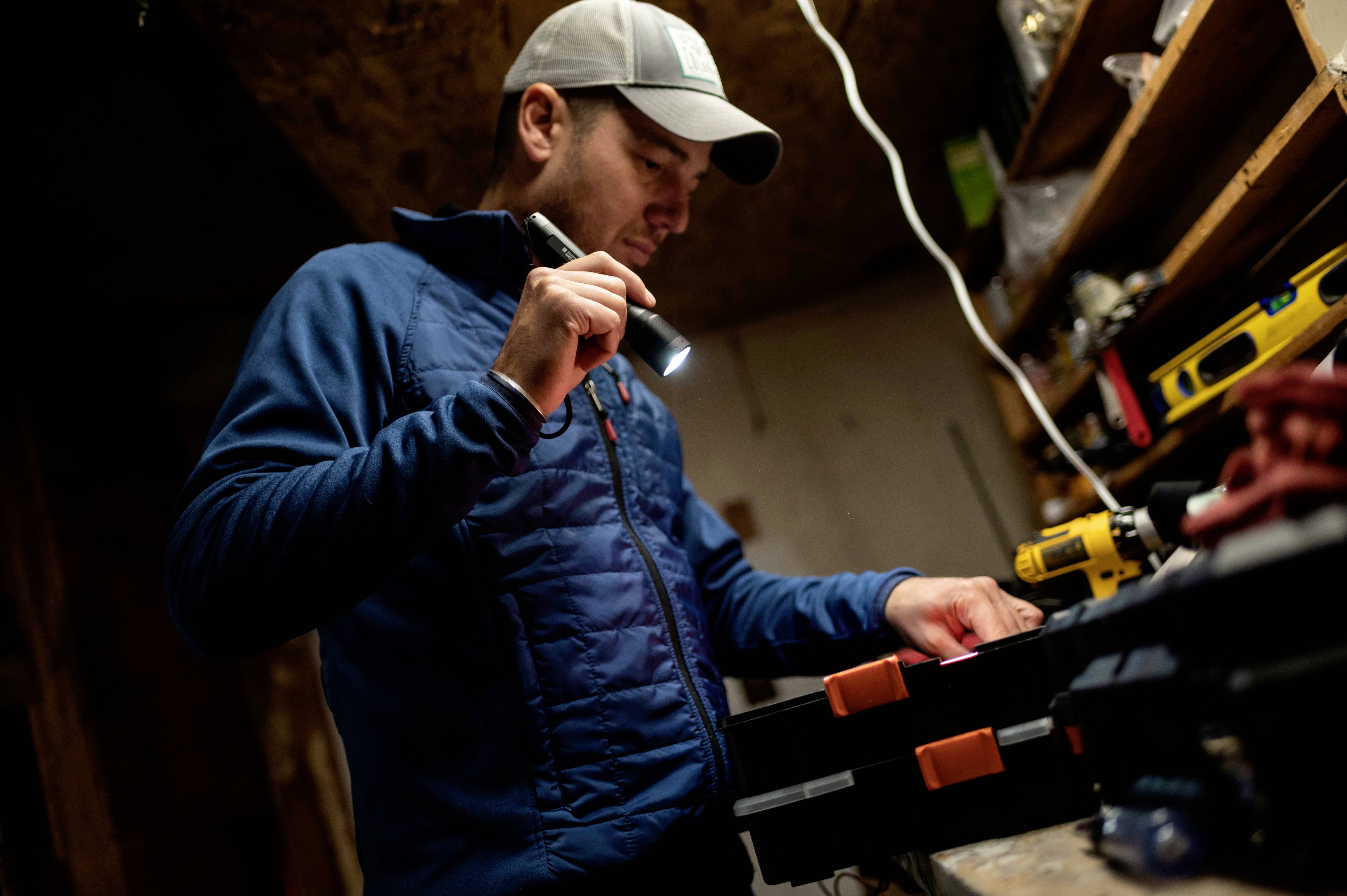 Un homme travaille dans un atelier et tient une lampe de poche. Il porte une casquette et une veste bleue, entouré d'outils.