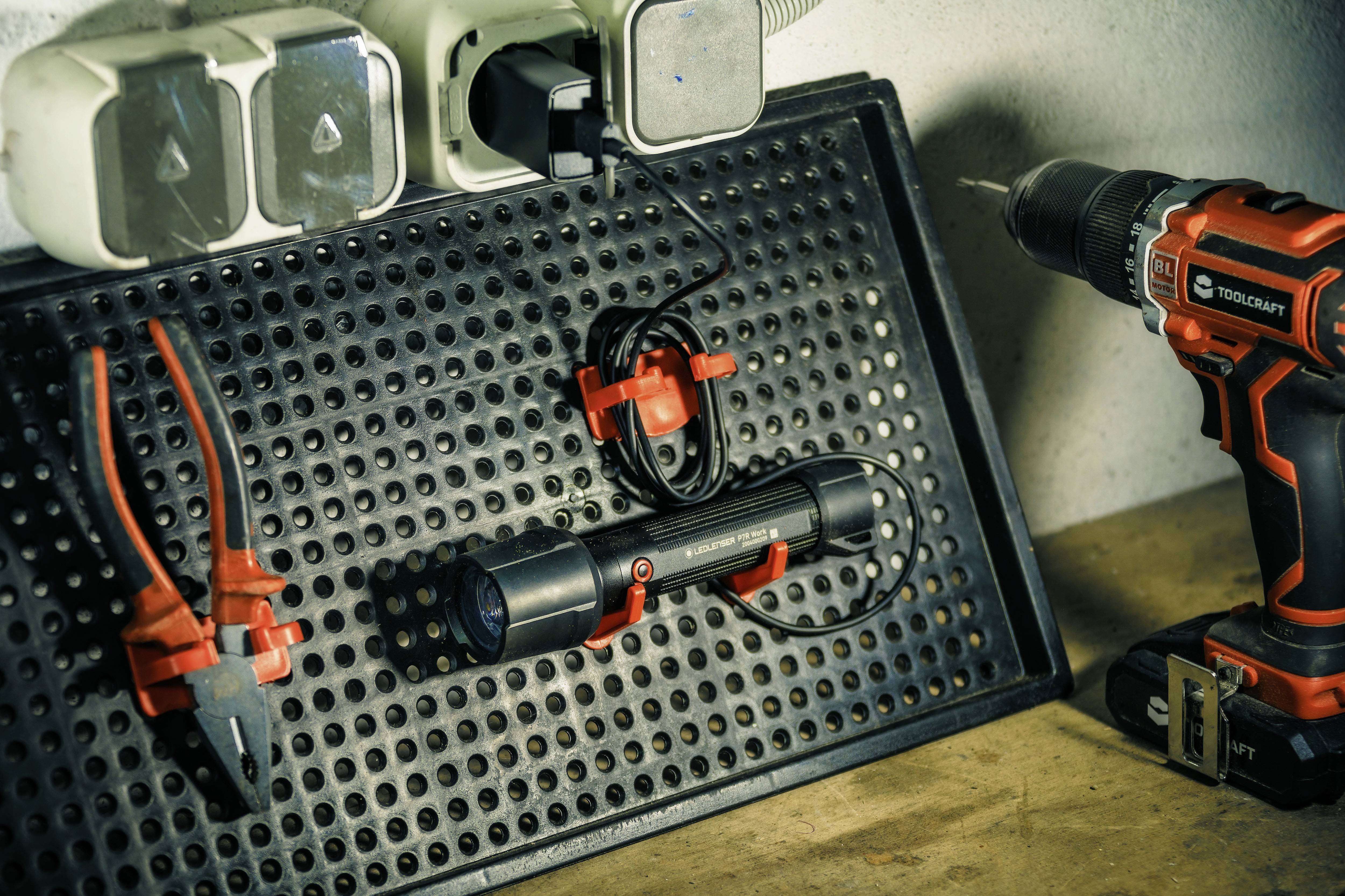 Sur un établi sont posés une lampe de poche noire, une pince rouge et une perceuse. Au fond, on aperçoit des prises électriques.