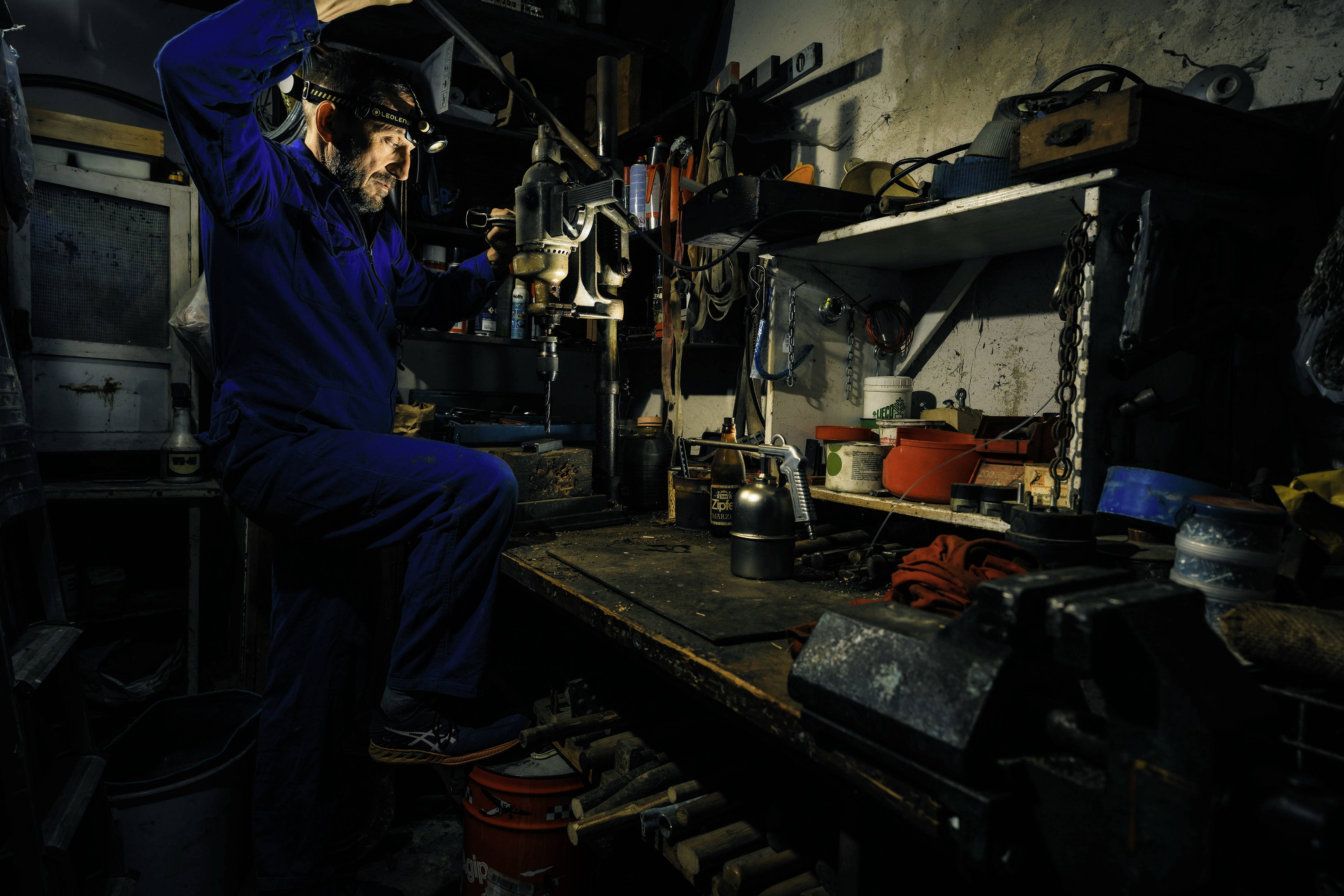 Artisan utilisant une perceuse dans un atelier. Environnement sombre et confiné avec des outils et des matériaux, un éclairage de travail dirigé sur la scène.