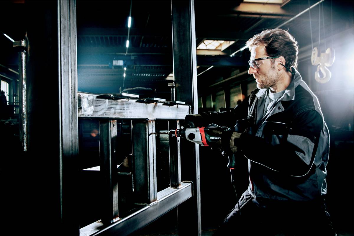 Un homme en tenue de protection travaille avec concentration sur un bâti métallique à l'aide d'une perceuse dans un environnement industriel d'atelier.