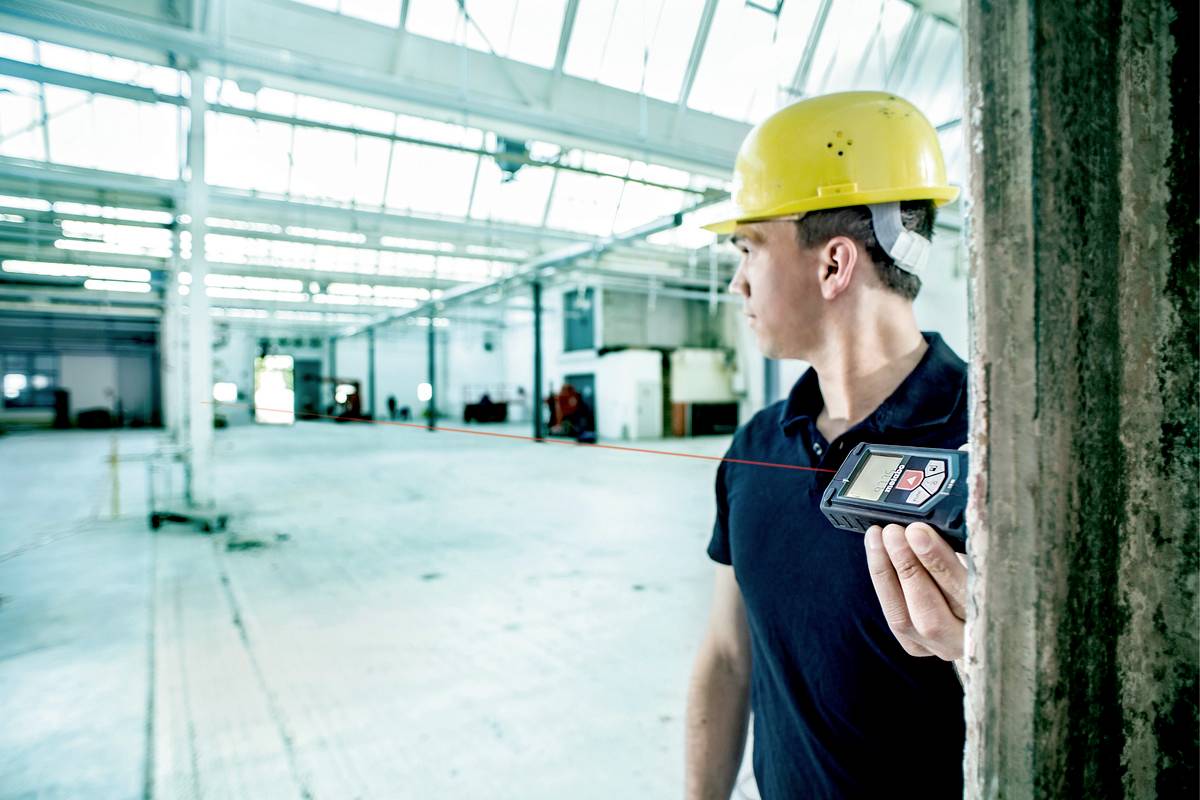 Un homme portant un casque de protection utilise un télémètre laser pour mesurer les dimensions d'un grand entrepôt vide.