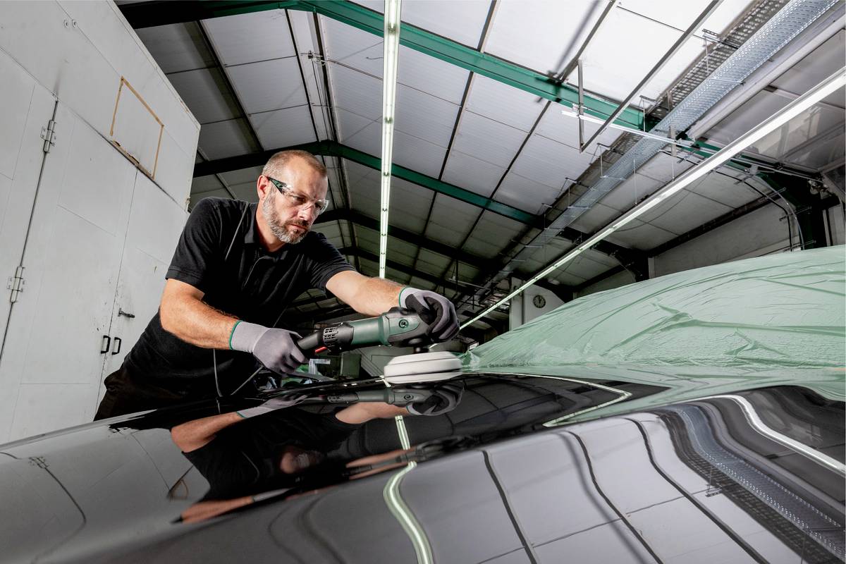Un homme polit le toit d'une voiture dans un atelier à l'aide d'une polisseuse. Il porte des lunettes de protection et des gants.