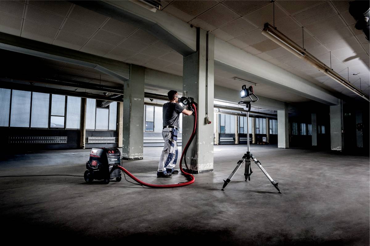 Une personne travaille dans un grand espace vide avec un aspirateur industriel et une source lumineuse sur un trépied.