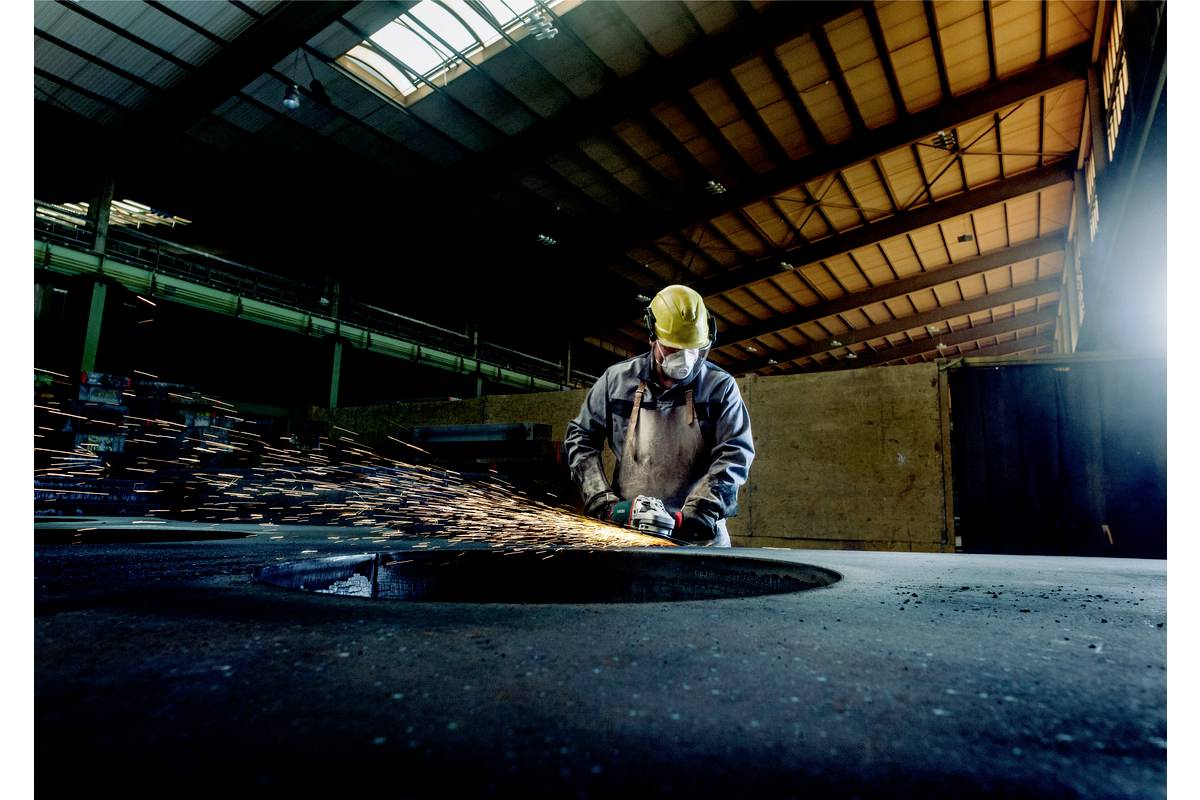 Un ouvrier en tenue de protection meule du métal dans un hall d'usine. Des étincelles jaillissent de la meuleuse. La lumière s'infiltre par les lanterneaux.