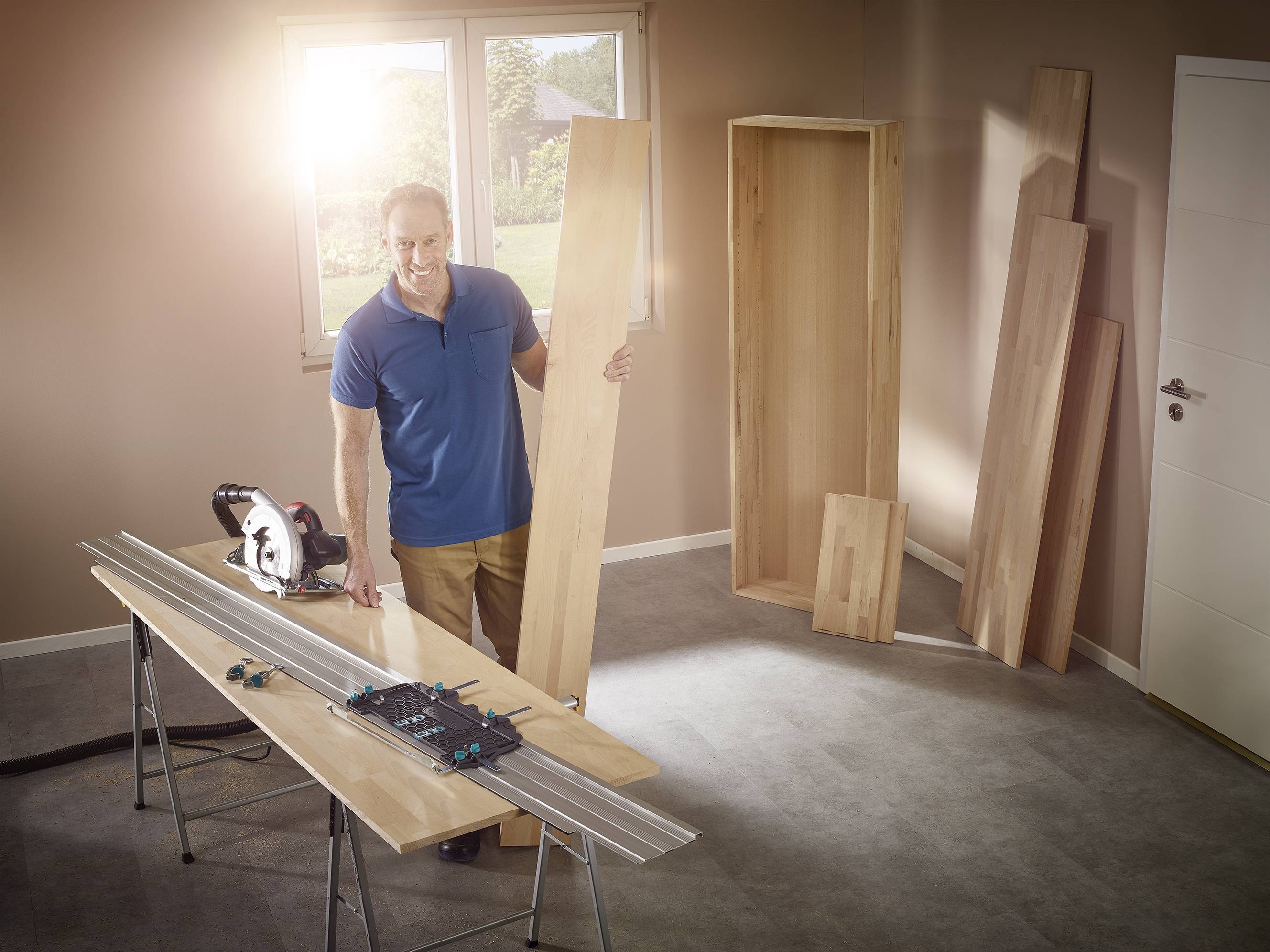 Un homme assemble une étagère en planches de bois dans une pièce lumineuse. Une scie circulaire est posée sur la table.