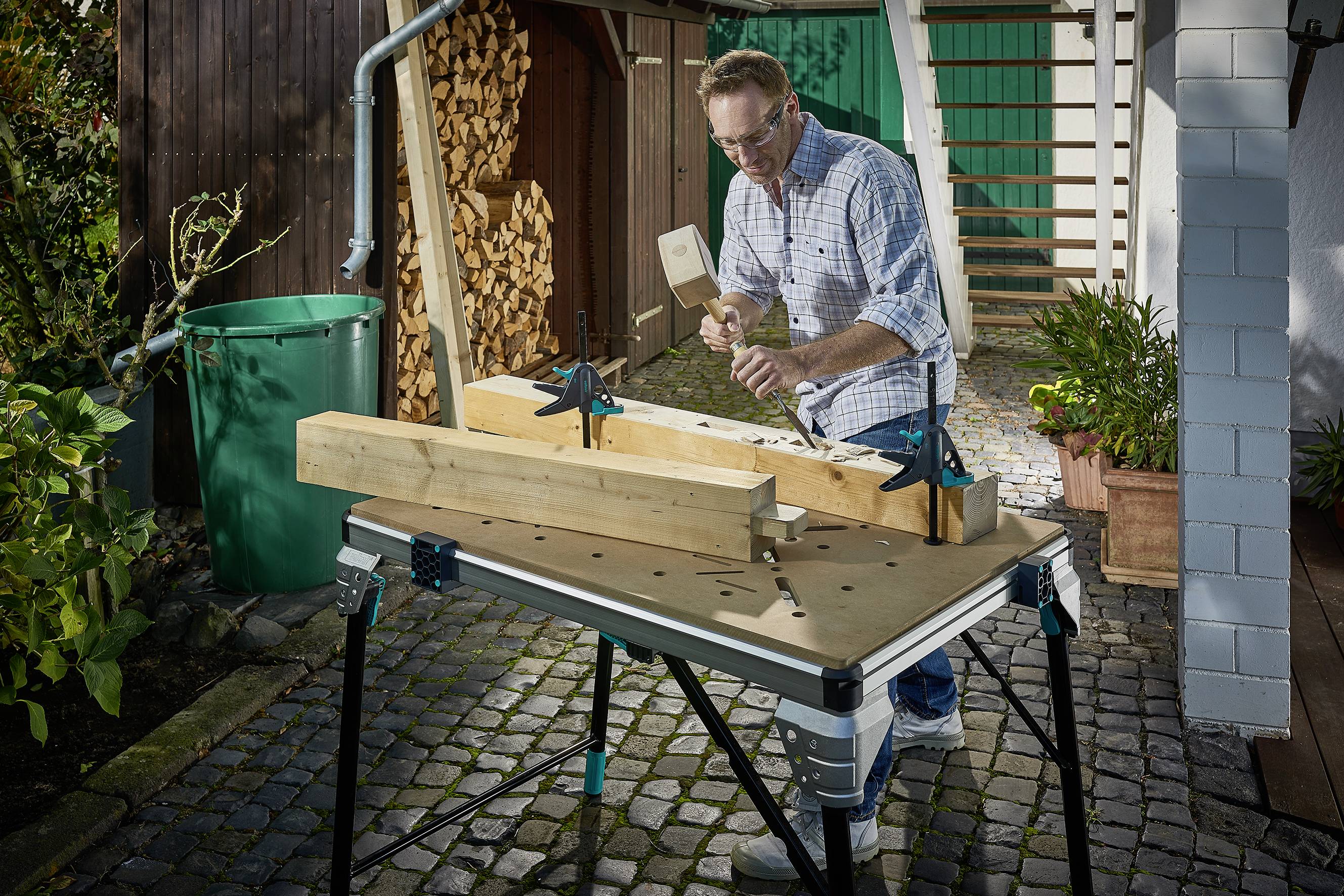 Un homme travaille un morceau de bois sur un établi à l'extérieur, entouré de plantes et de piles de bois, portant des lunettes de protection et utilisant un marteau.