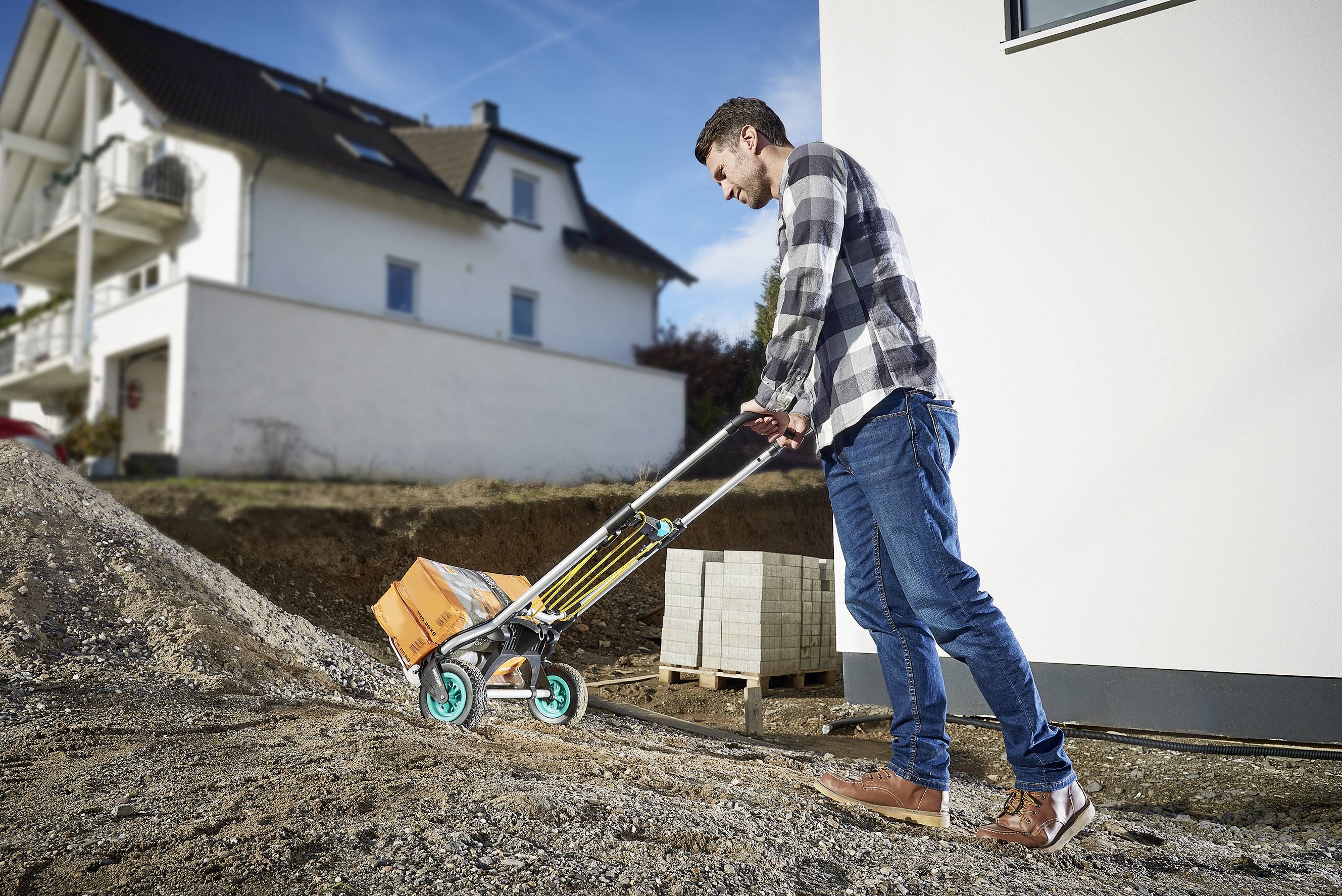 Un homme pousse un sac de ciment sur une brouette le long d'une colline. À l'arrière-plan, on aperçoit une maison et un ciel bleu.