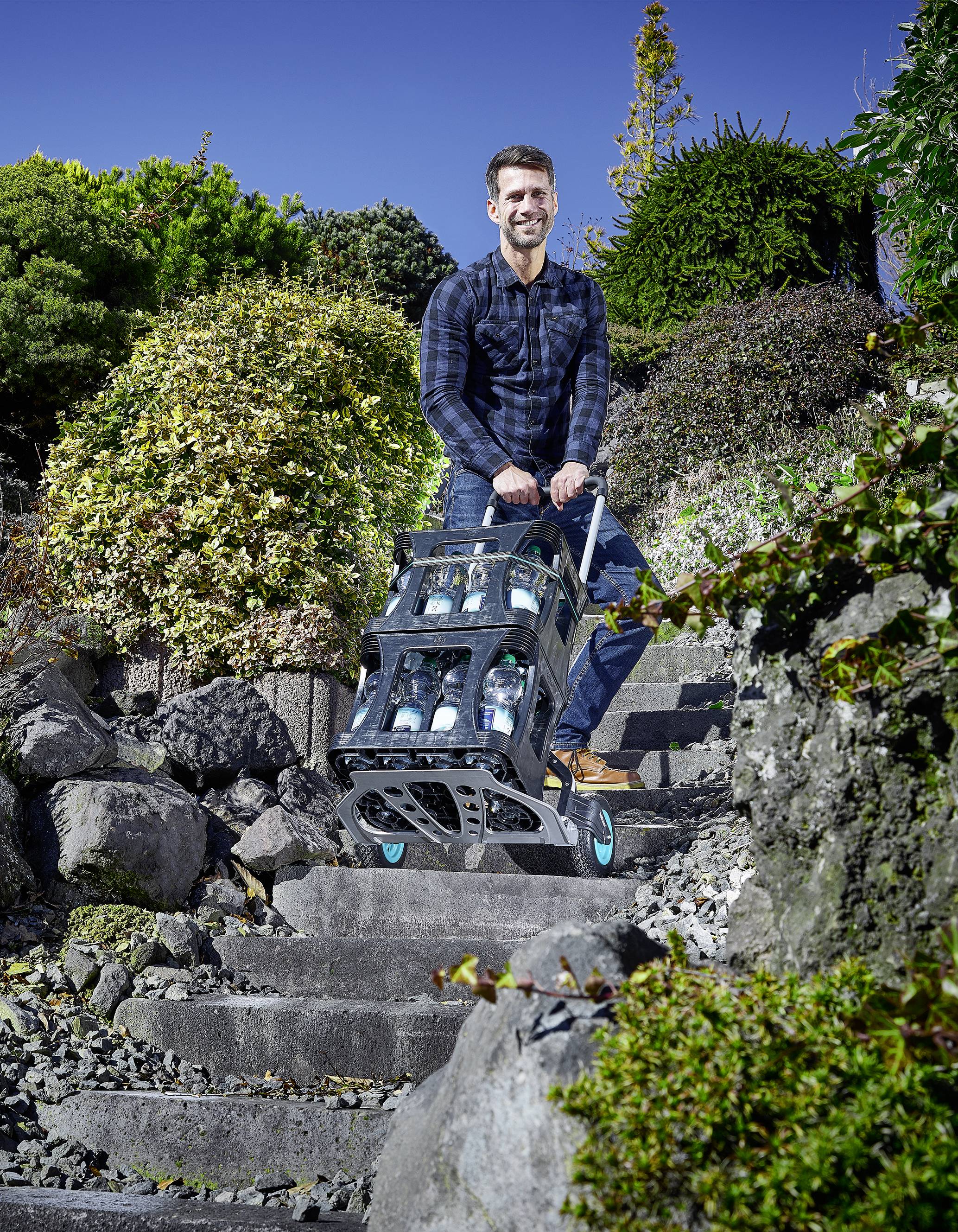 Un homme transporte plusieurs bouteilles d'eau sur un chariot à main le long d'un escalier en pierre. Des plantes sont visibles en arrière-plan.