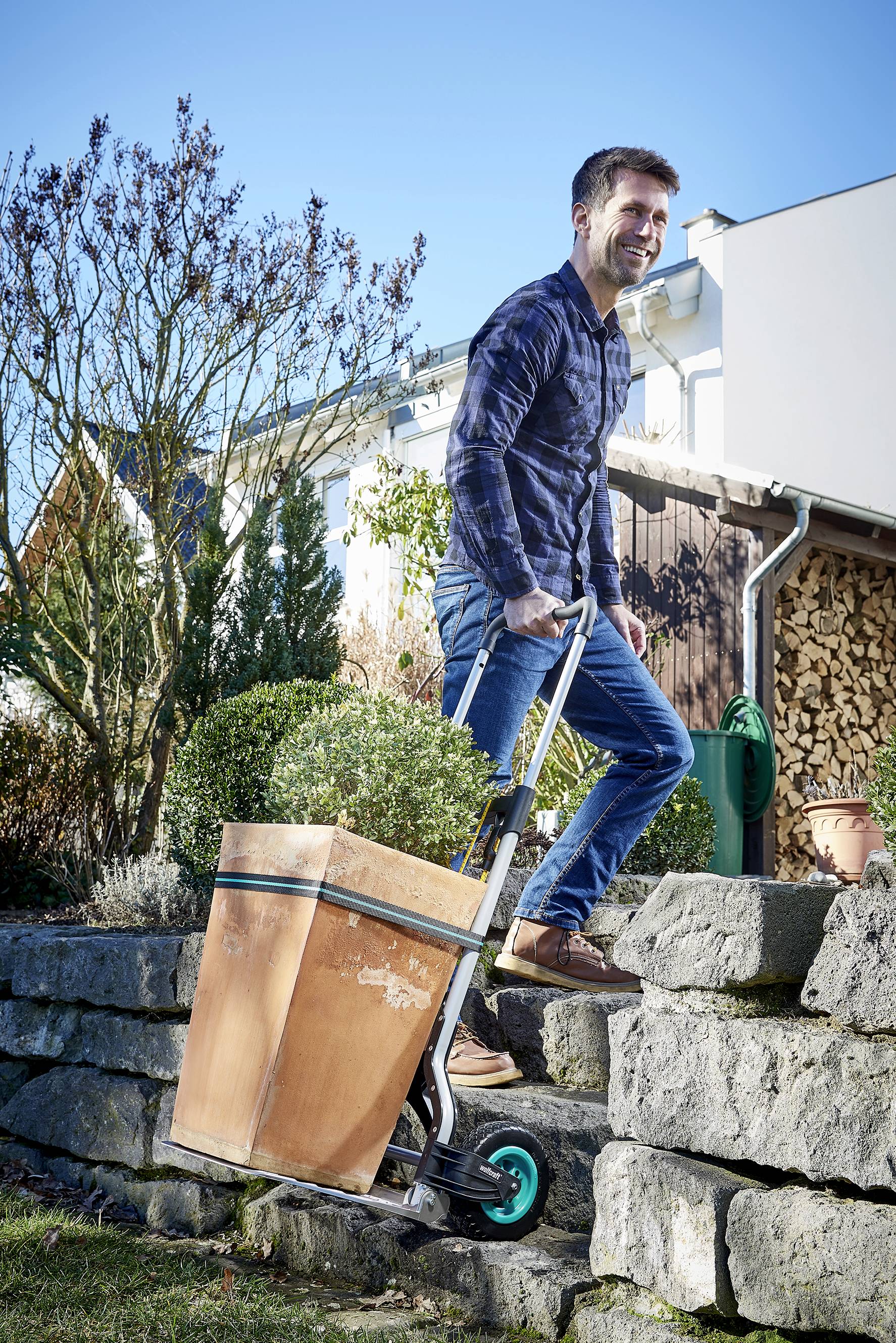 Un homme transporte un grand pot de fleurs avec un diable le long d'un escalier en pierre dans un jardin par une journée ensoleillée.