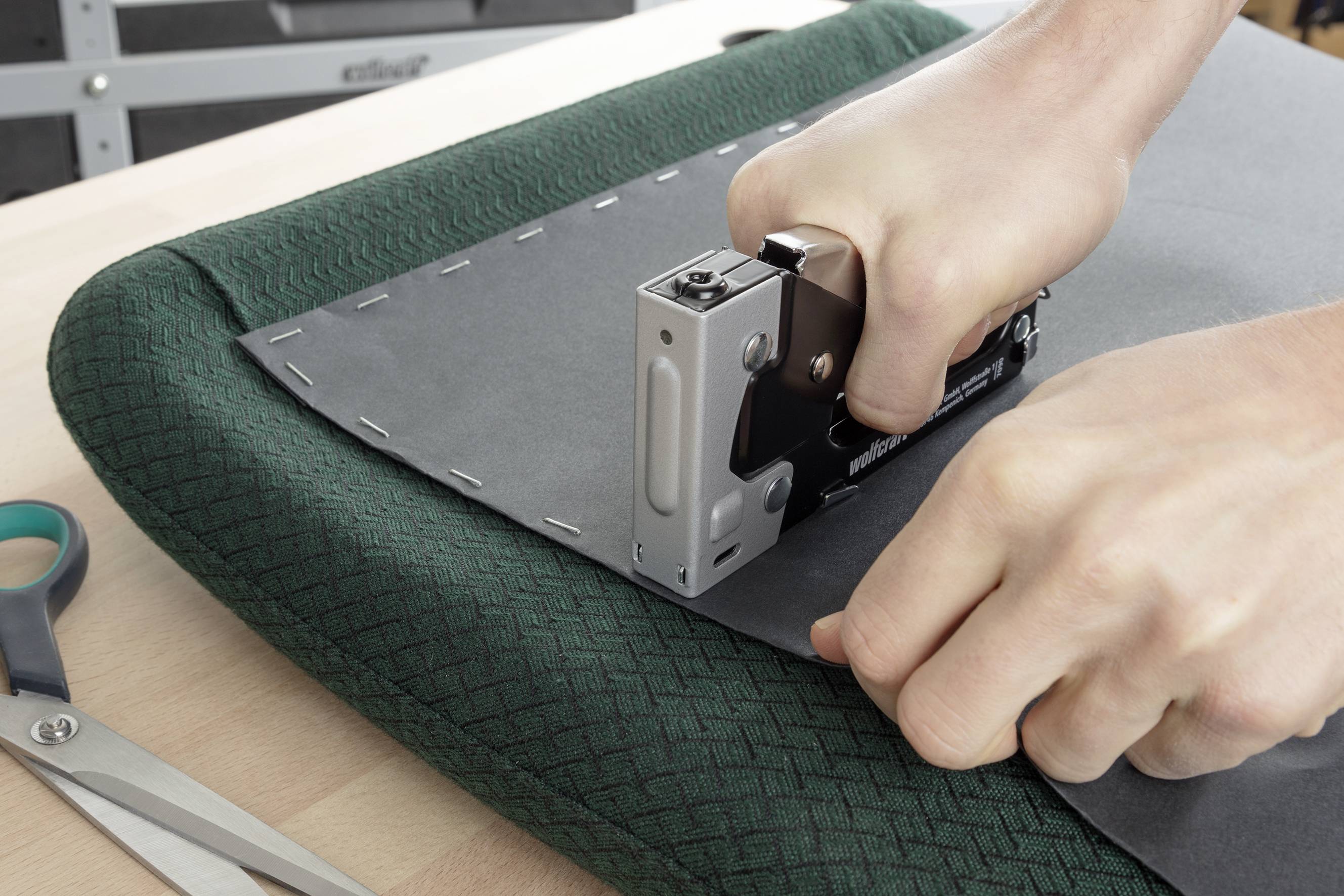 Une personne fixe une housse grise en tissu sur un coussin à l'aide d'un pistolet à agrafes. Une paire de ciseaux est posée à côté sur la table.