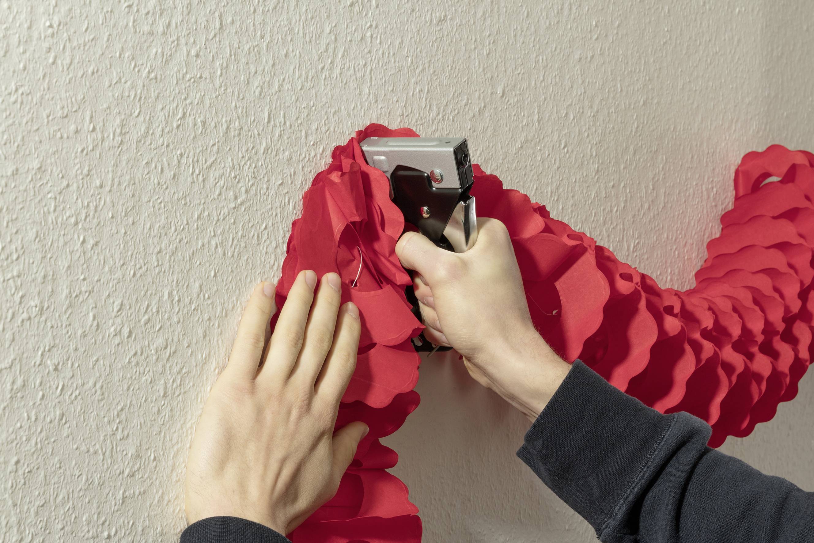 Une personne fixe une décoration rouge au mur avec un pistolet à agrafes. Les mains maintiennent fermement la décoration.