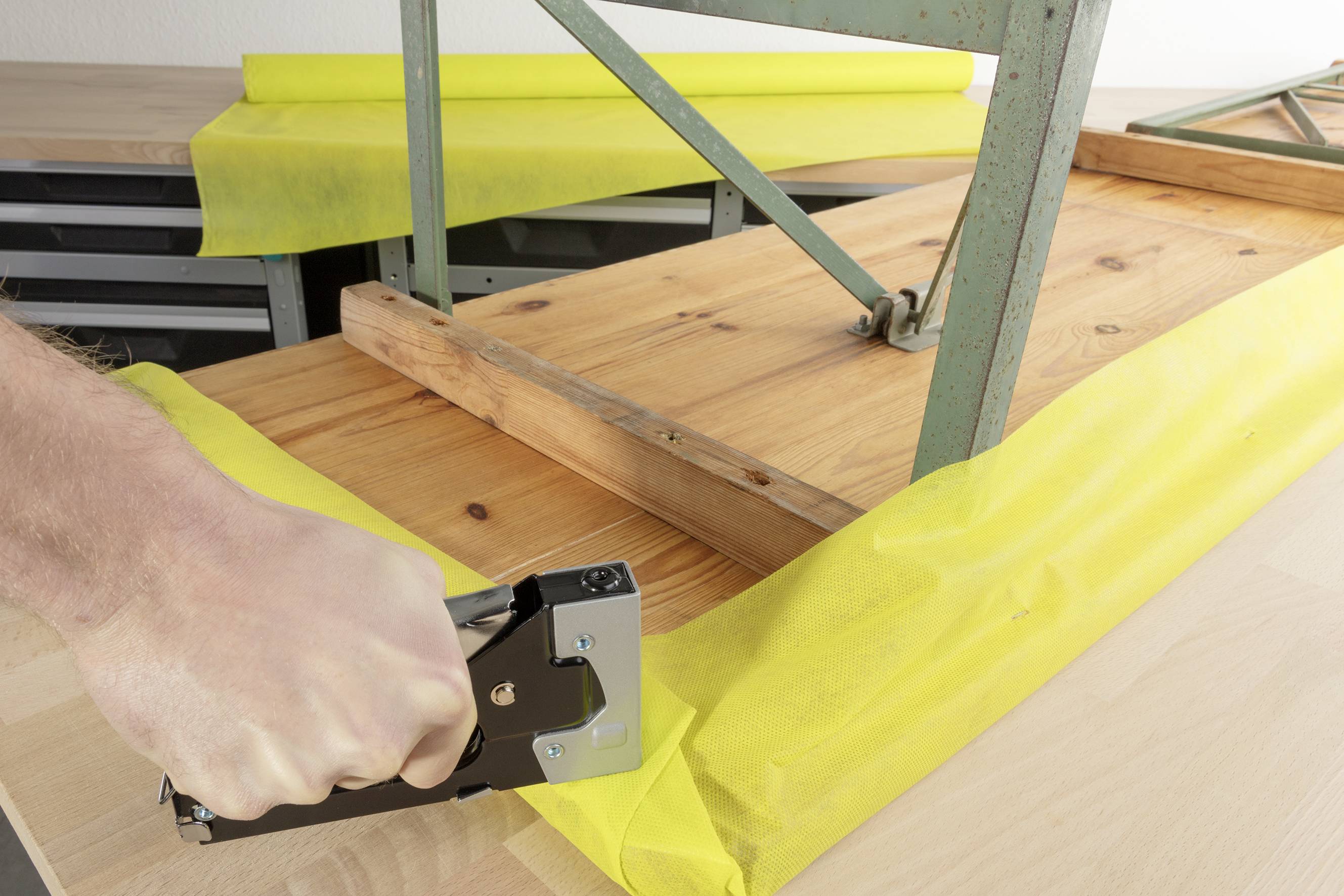 Une main utilise un pistolet à agrafes pour fixer un tissu jaune à une table en bois. Un cadre métallique est posé sur la table.