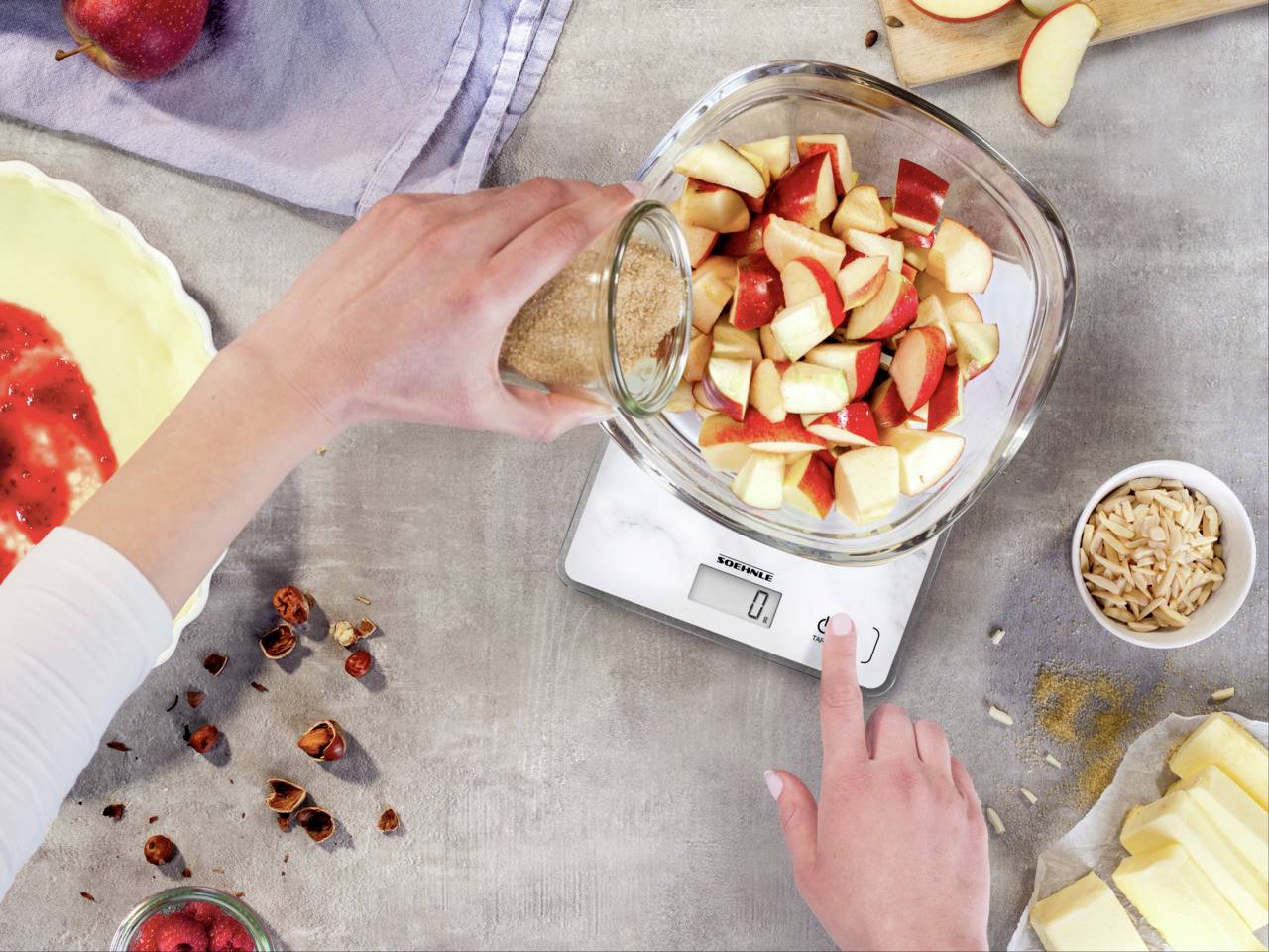 Une main pèse des morceaux de pomme dans un bol en verre sur une balance de cuisine, à côté d'ingrédients tels que des noix et du beurre sur une table de cuisine.