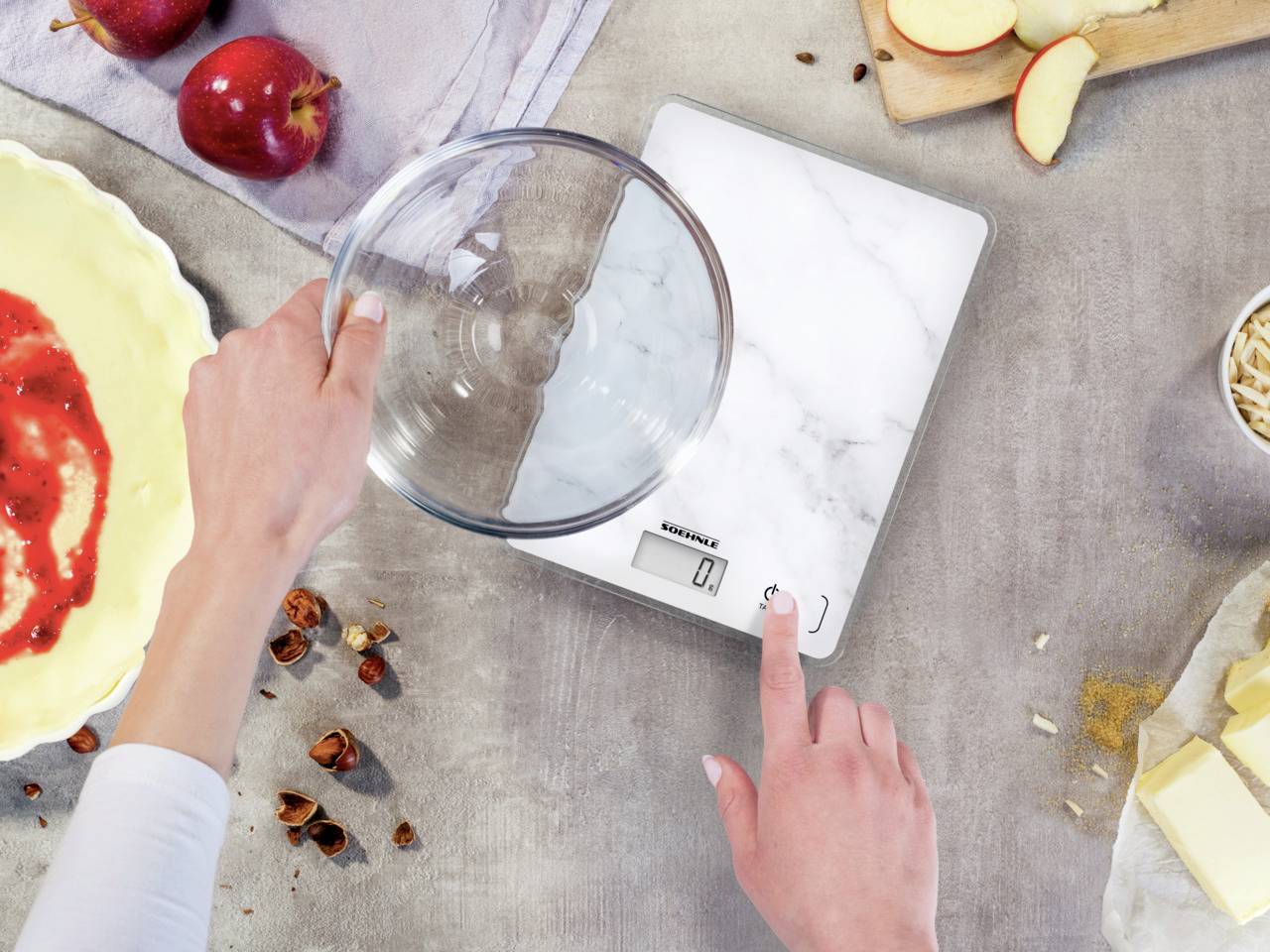 Une personne pèse un bol à mélanger vide sur une balance de cuisine digitale, entourée d'ingrédients tels que des pommes, des noix et du beurre pour un gâteau.