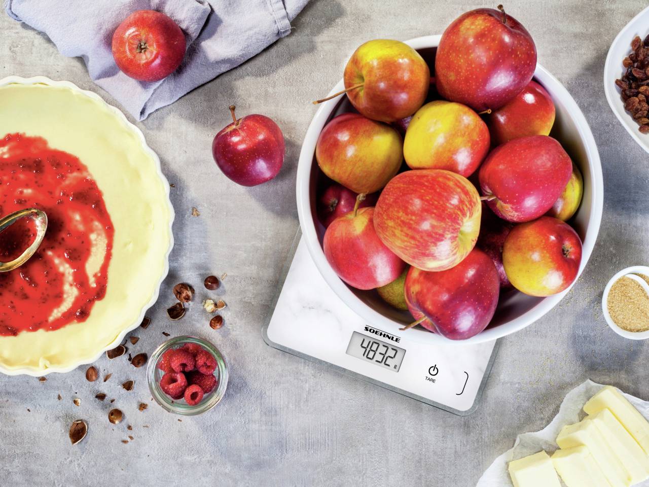 Bol de pommes sur une balance (poids : 483,2 g), à côté une pâte à gâteau avec une garniture rouge et des baies, préparation pour une tarte aux pommes.