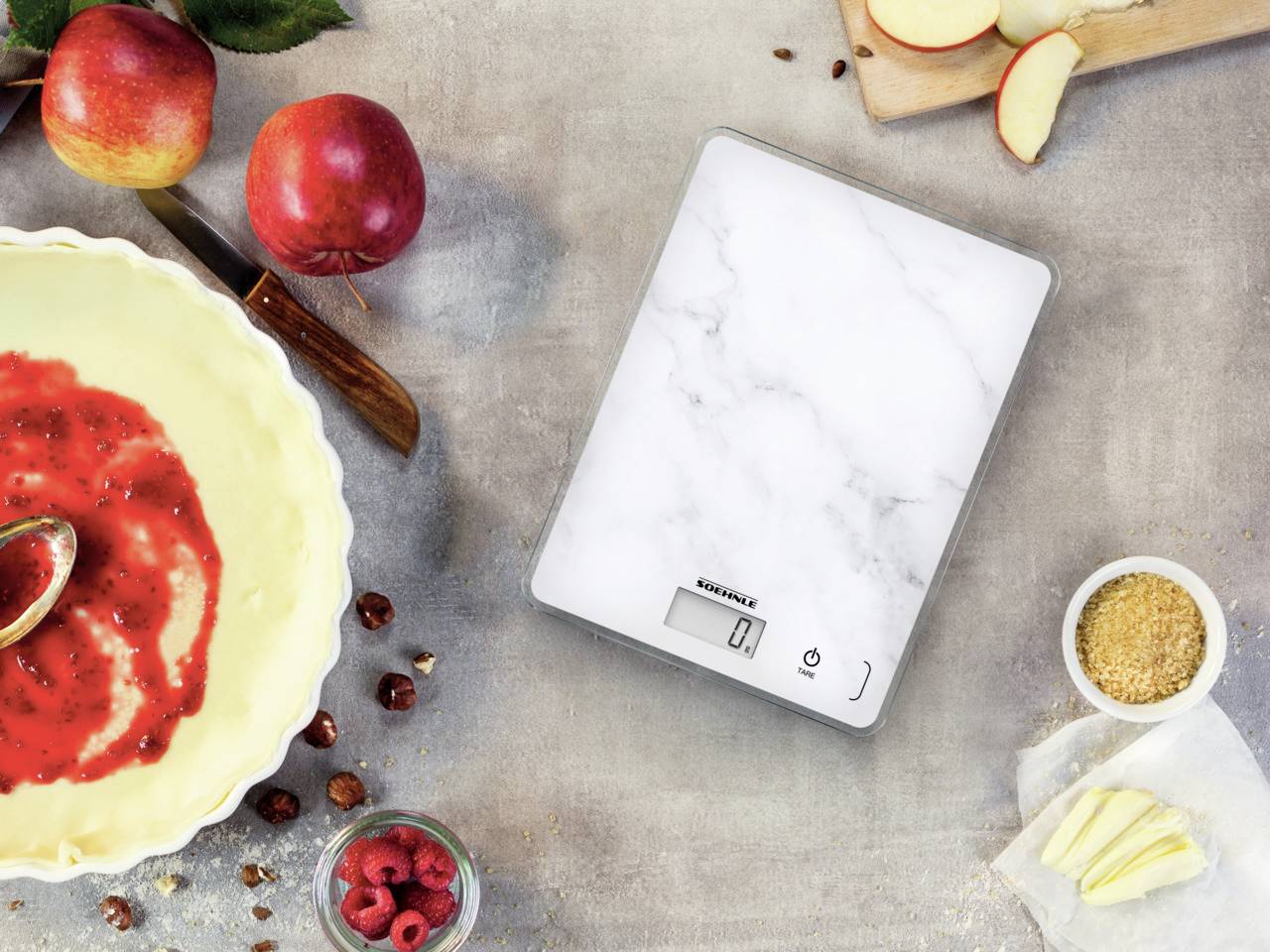Balance numérique de cuisine posée sur une table, entourée d'ingrédients tels que des pommes, des framboises, des noisettes et une sauce aux fruits rouges dans un bol.