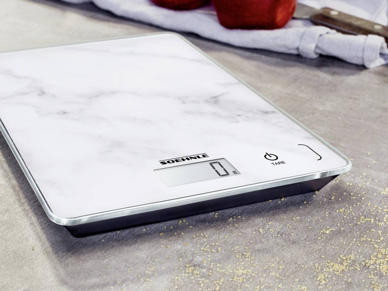 Balance de cuisine moderne sur une surface en marbre affichant '0 g' sur son écran numérique, entourée de pommes et d'un torchon.