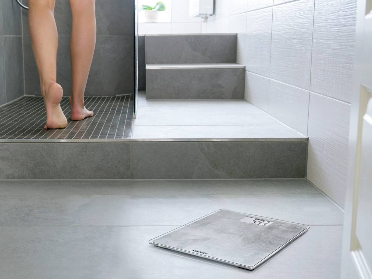 Une personne quitte la douche avec un pèse-personne numérique sur le sol. L'environnement montre une salle de bain carrelée avec des marches et des carreaux gris modernes.
