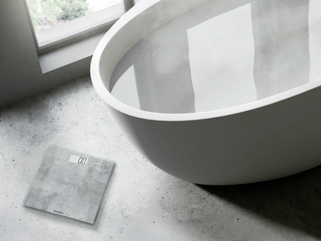 Une balance est placée à côté d'une baignoire blanche moderne sur un sol gris, éclairée par la lumière naturelle.