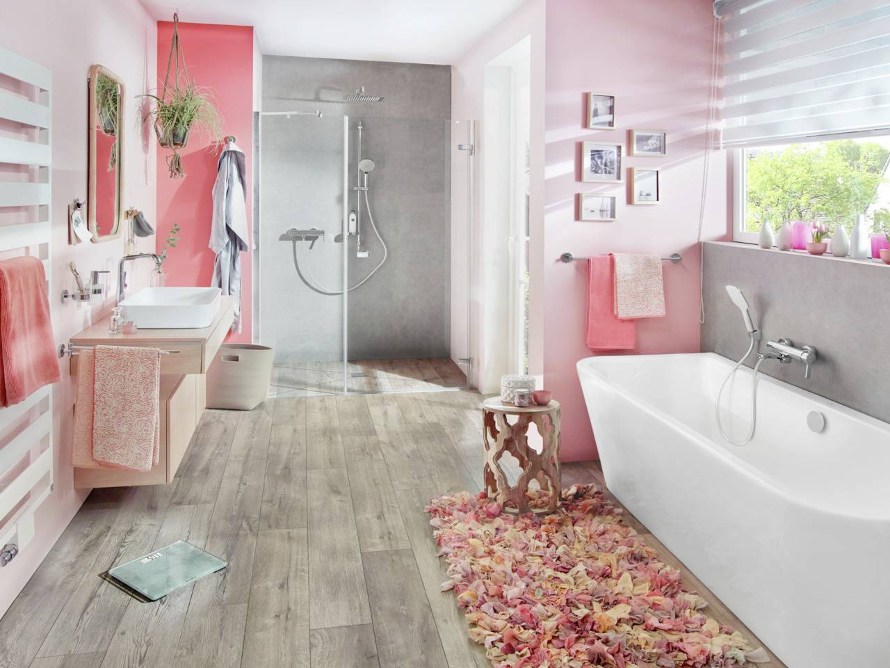 Salle de bain moderne avec des touches de rose, baignoire îlot, douche à effet pluie et sol en bois. Des décorations murales et des plantes complètent l'espace.