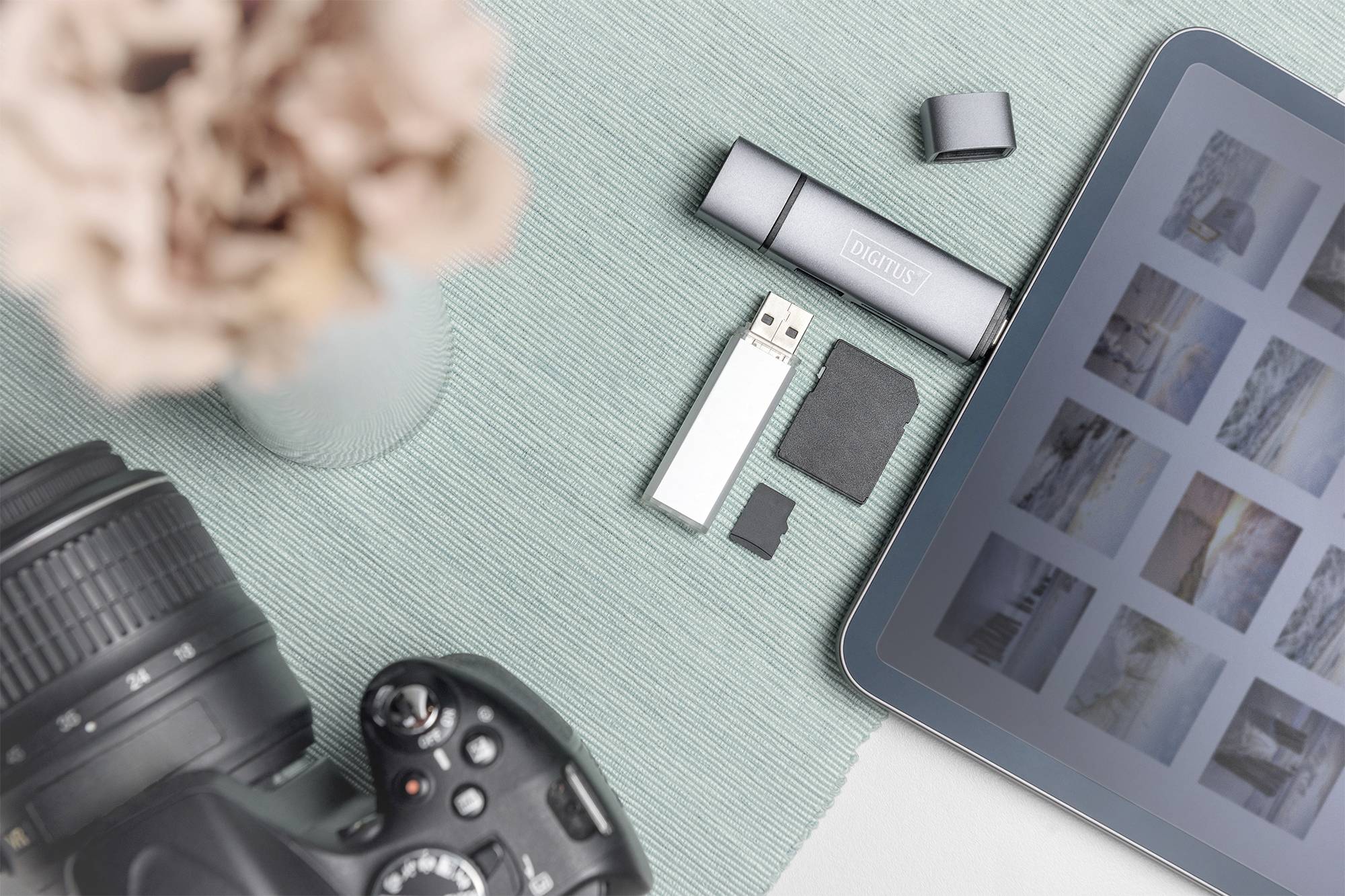 Un appareil photo reflex, une tablette avec une galerie photo, une clé USB et des cartes SD sur un support turquoise, à côté de fleurs.