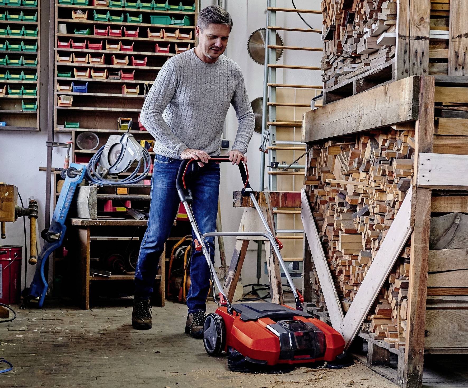 Un homme utilise un balai mécanique dans un atelier pour nettoyer le sol près d'une pile de bois. Des outils sont visibles en arrière-plan.