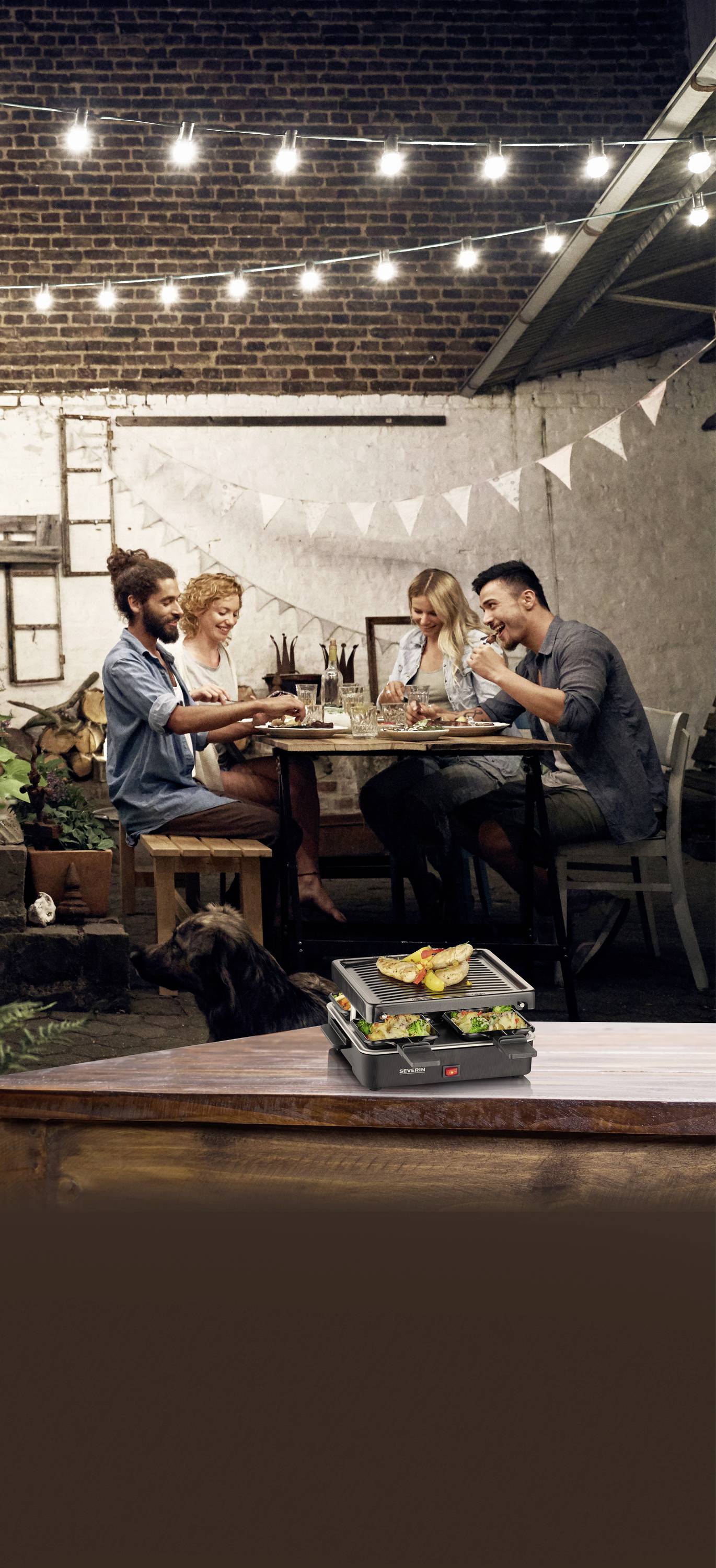 Un groupe de quatre personnes est assis à une table en extérieur, mange ensemble et discute. Au-dessus d'eux, des guirlandes lumineuses sont suspendues.